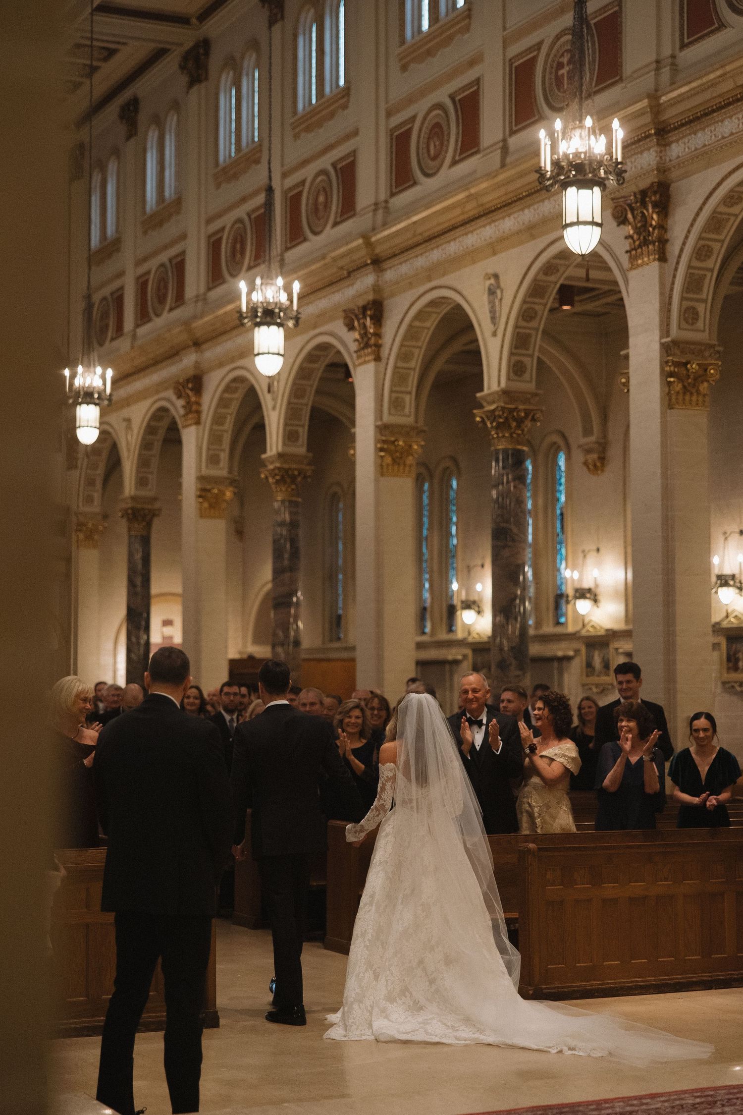 A traditional wedding ceremony takes place in an ornate church interior with grand arches and hanging chandeliers.