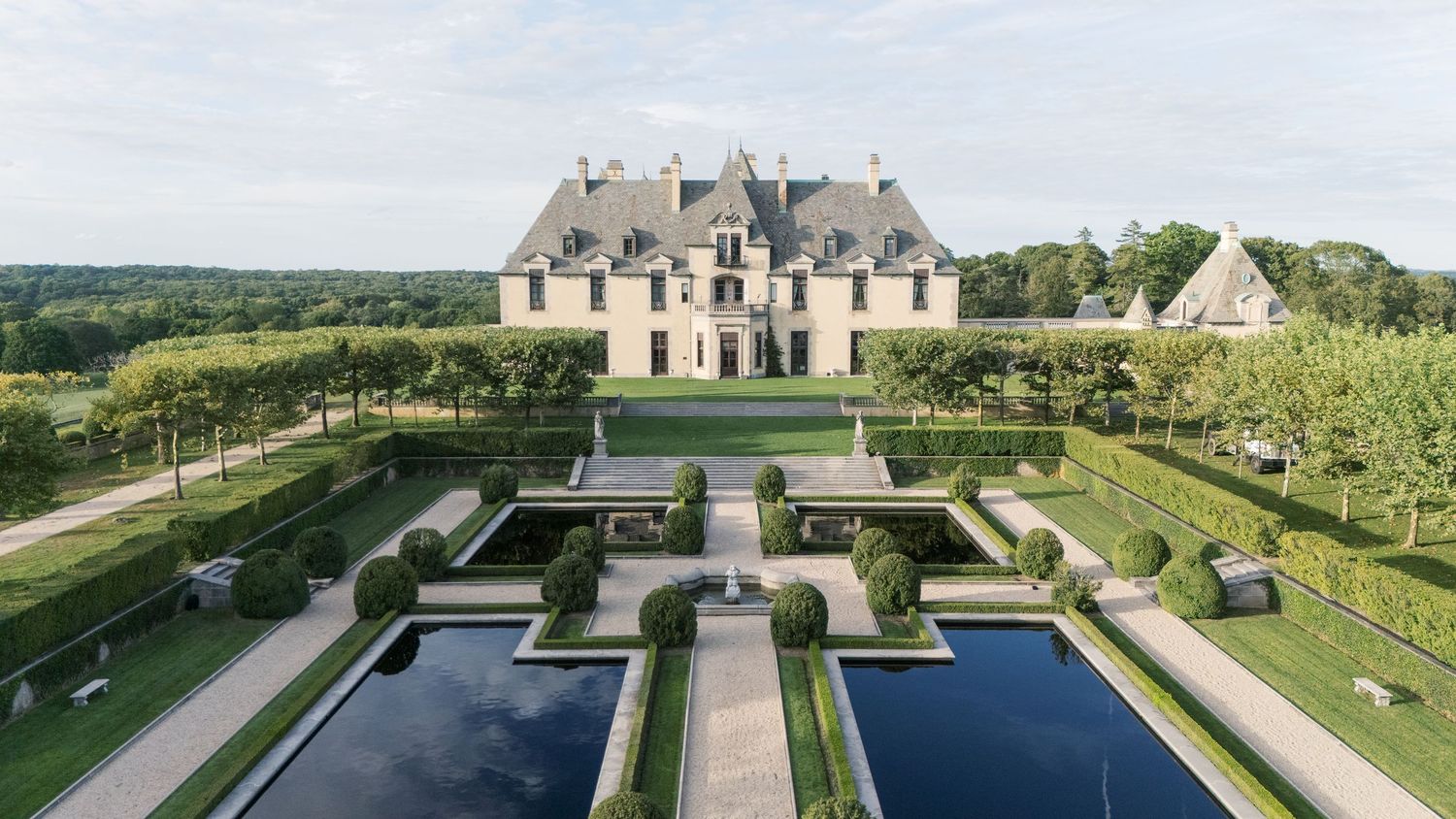 A panoramic aerial view shows a grand estate with manicured gardens, fountains and symmetrical landscaping in the French style.