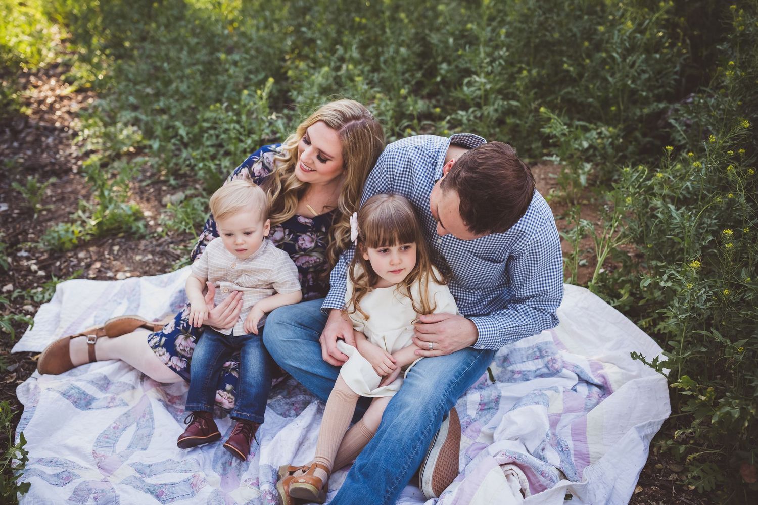A family sits together on a white blanket outdoors enjoying a sunny day in nature.