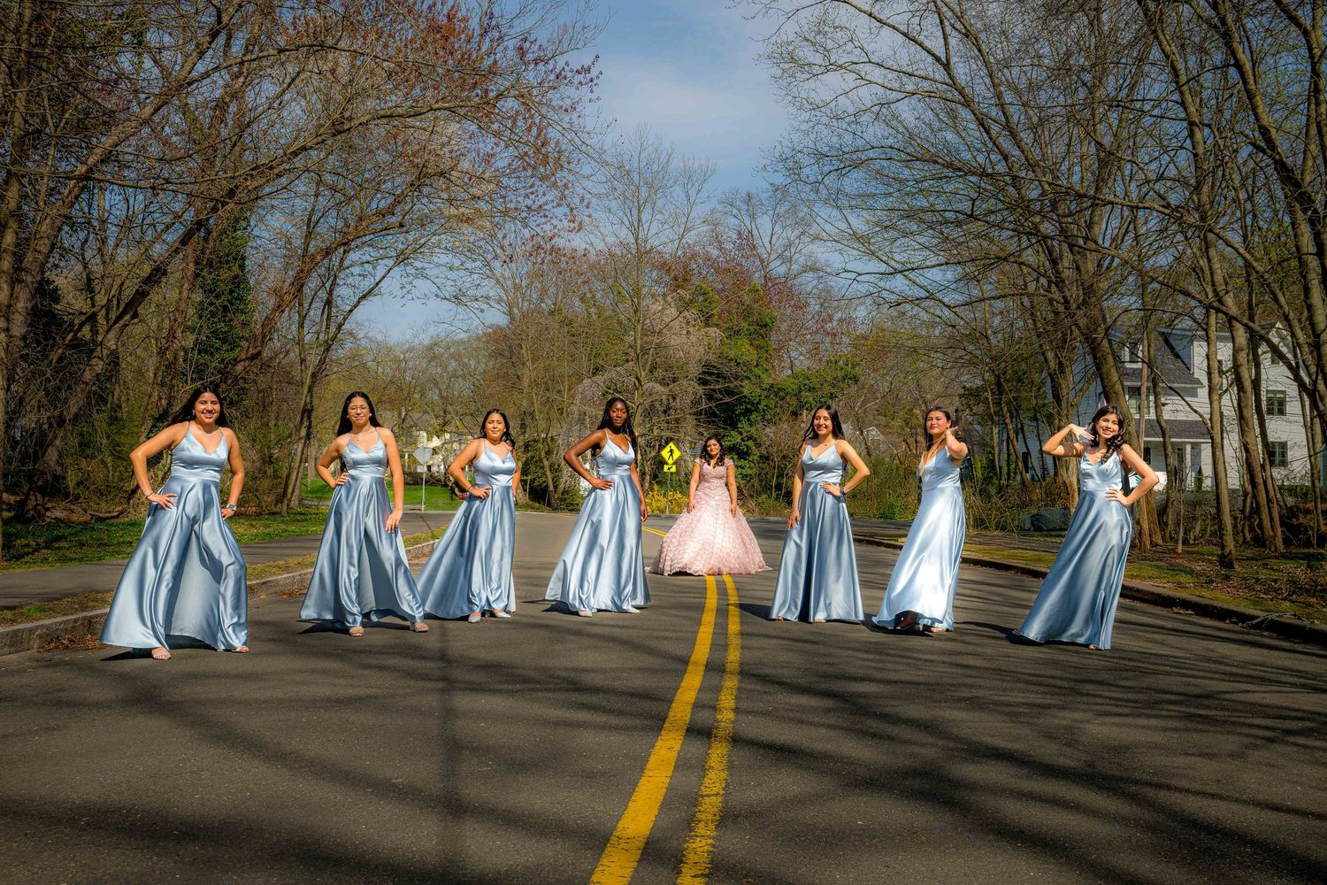 People in blue dresses stand in a line on a road surrounded by bare trees beneath a cloudy sky.