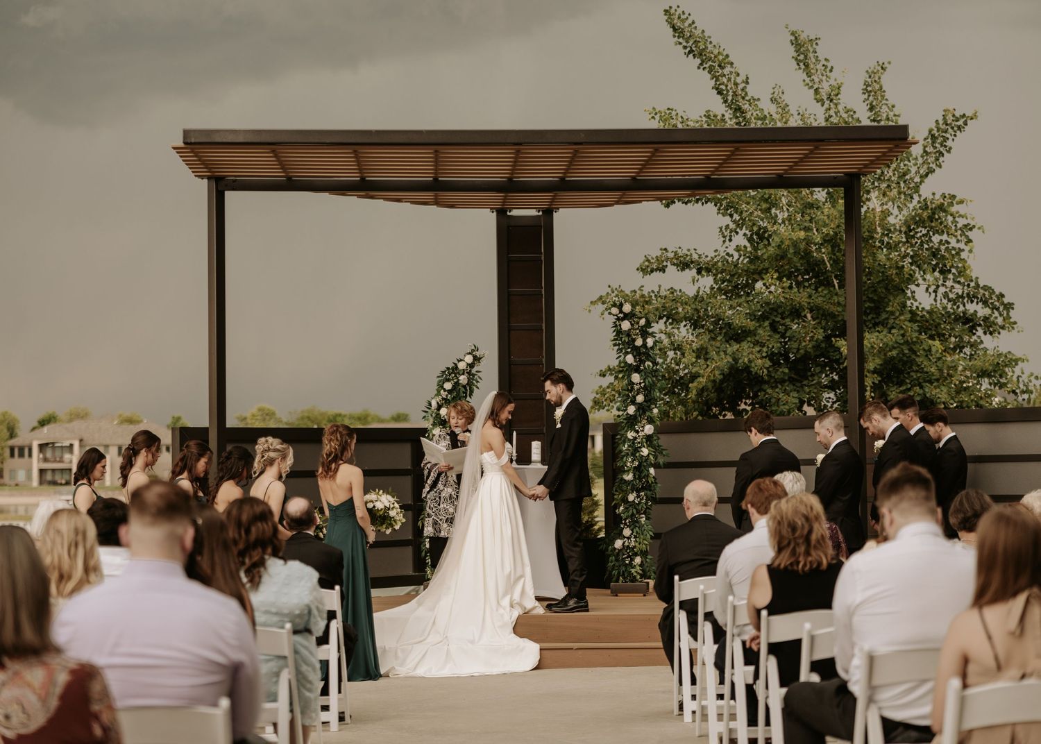 A wedding ceremony panorama shows guests seated in white chairs under an outdoor pergola structure at a modern venue.