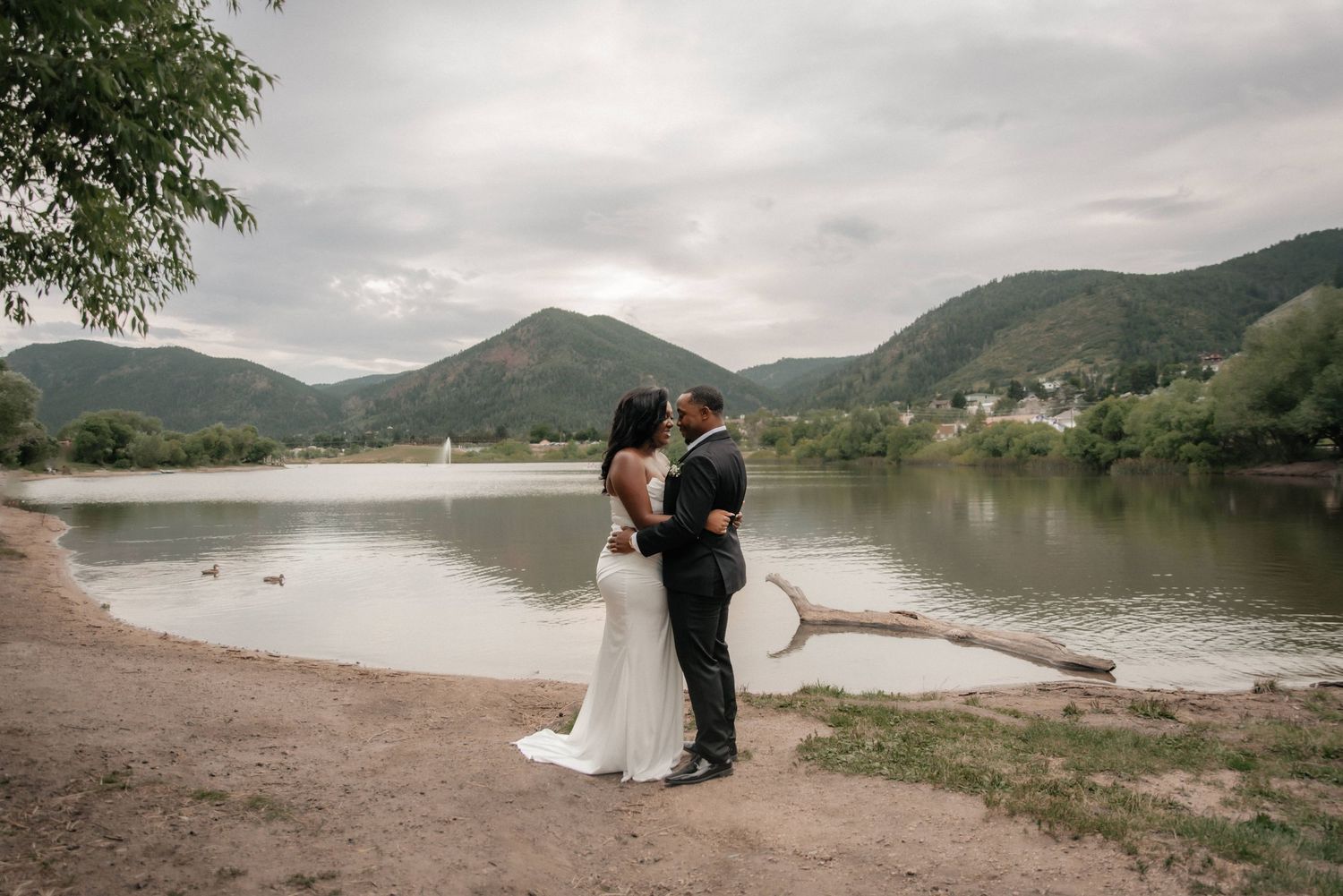 A couple shares a romantic moment by a serene mountain lake on an overcast day.