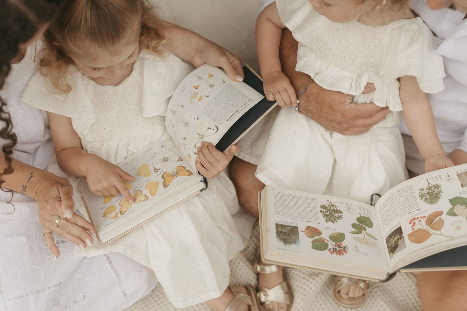 Children in white dresses look through books with floral illustrations while sitting together on a textured surface.