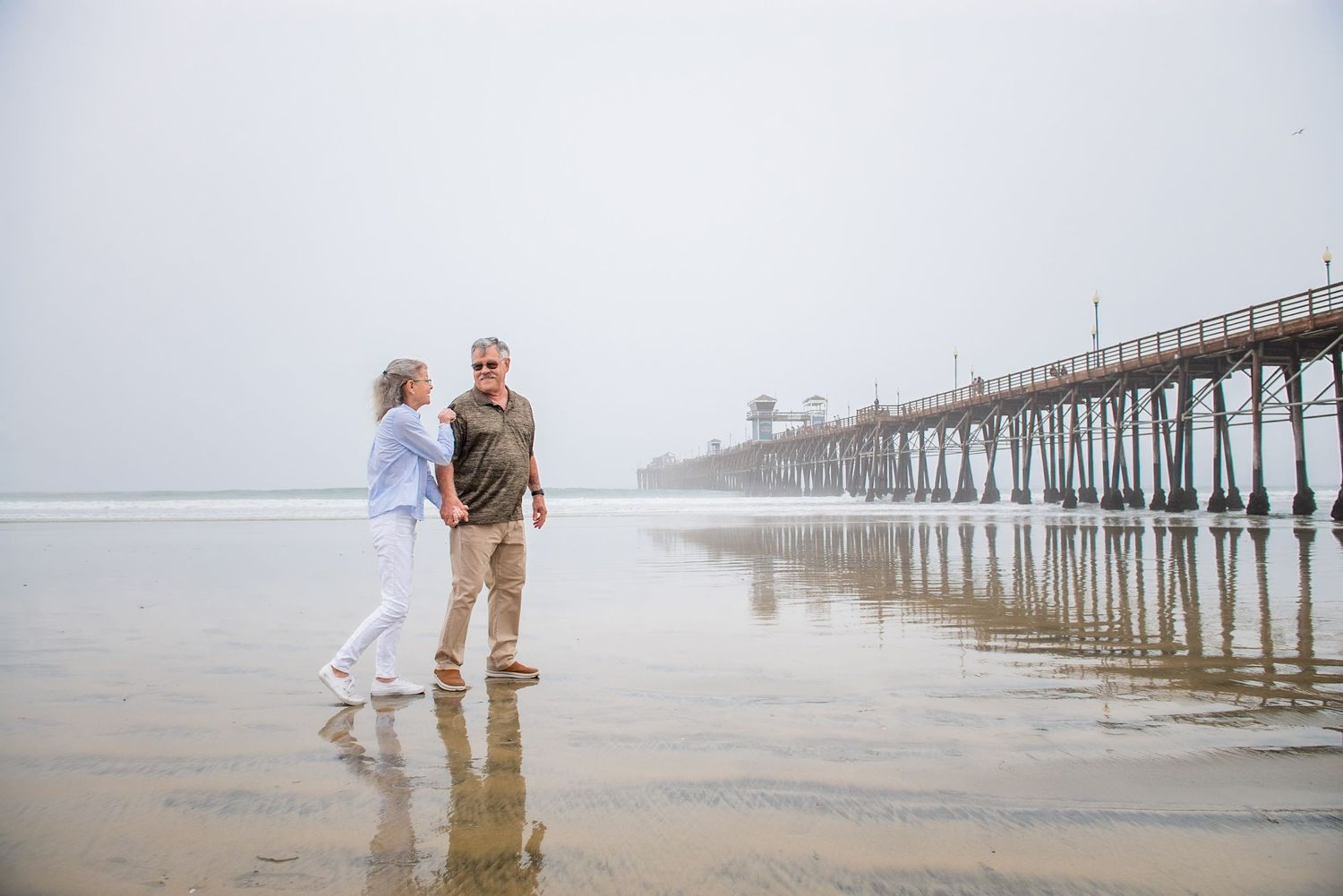 Two people stand together on misty beach near Oceanside Pier with reflections in wet sand during foggy morning.