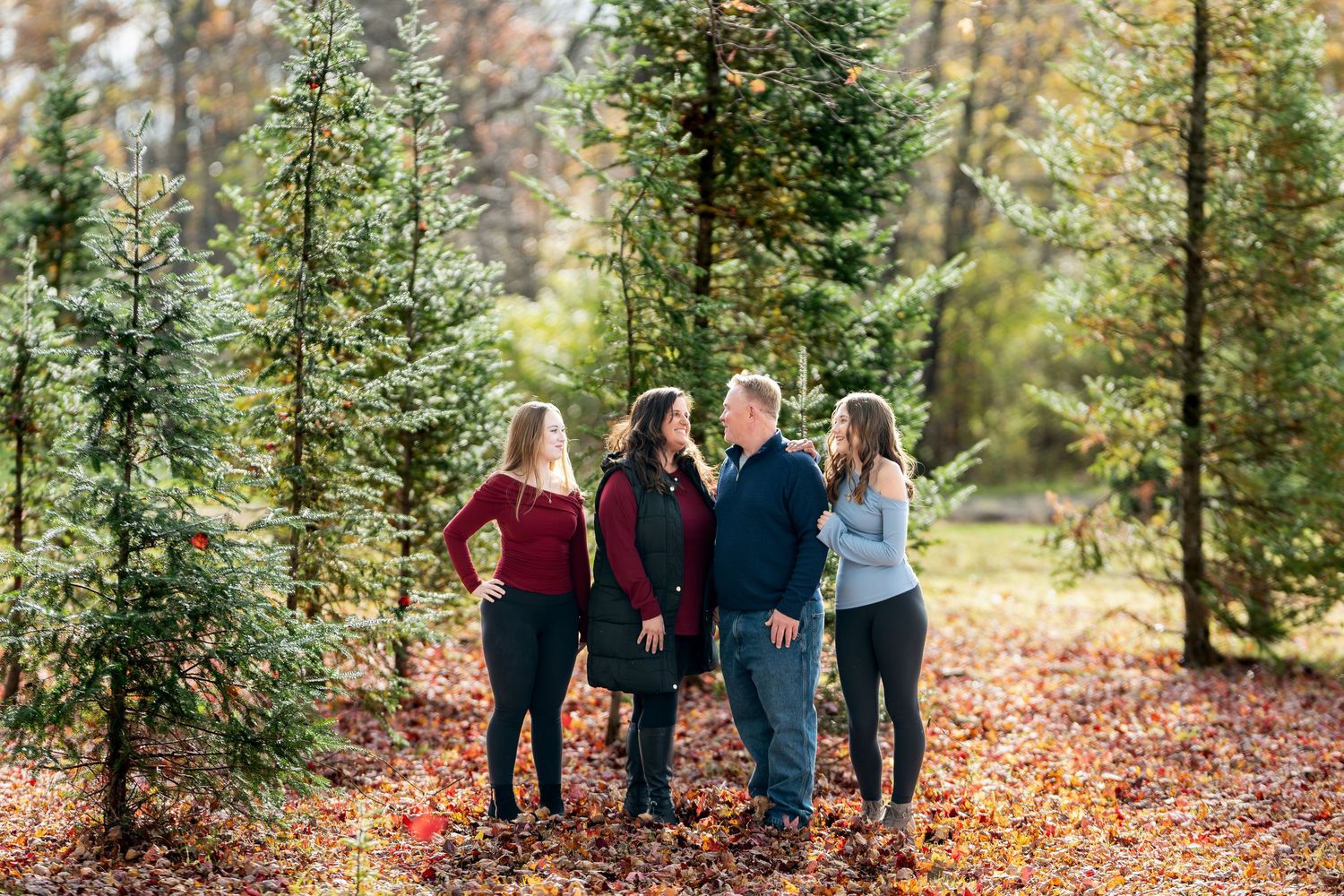 Family of four smiling together among evergreen trees in a colorful autumn Christmas tree farm setting.