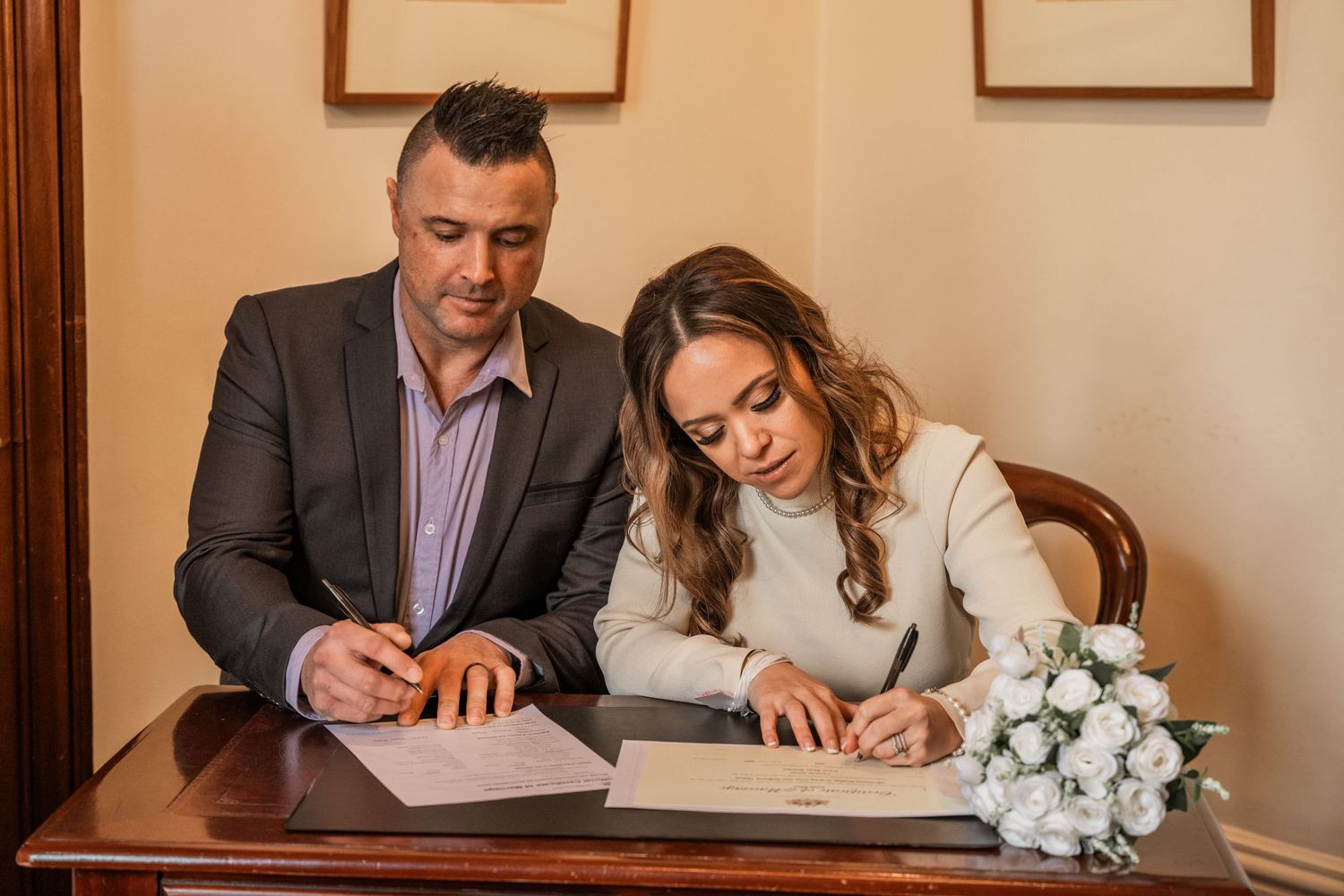 A bride and groom signing their marriage license with white flower bouquet on table.