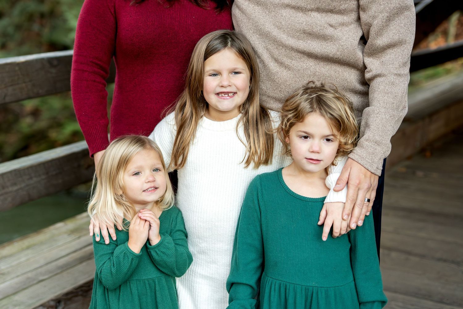 Three young girls in green and white dresses posing together on a wooden bridge for family photos.