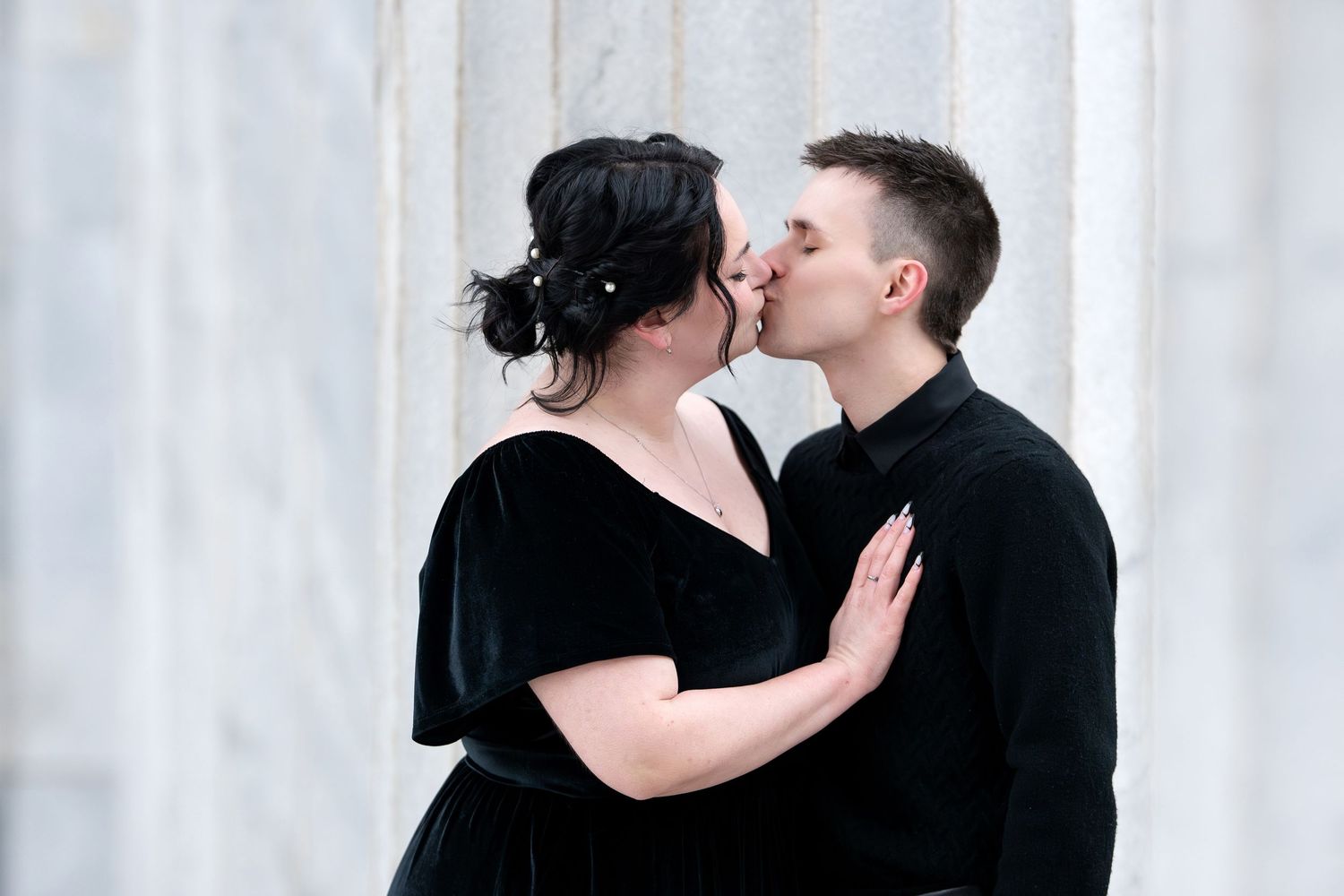 Couple sharing a tender kiss between marble columns, wearing formal black attire.
