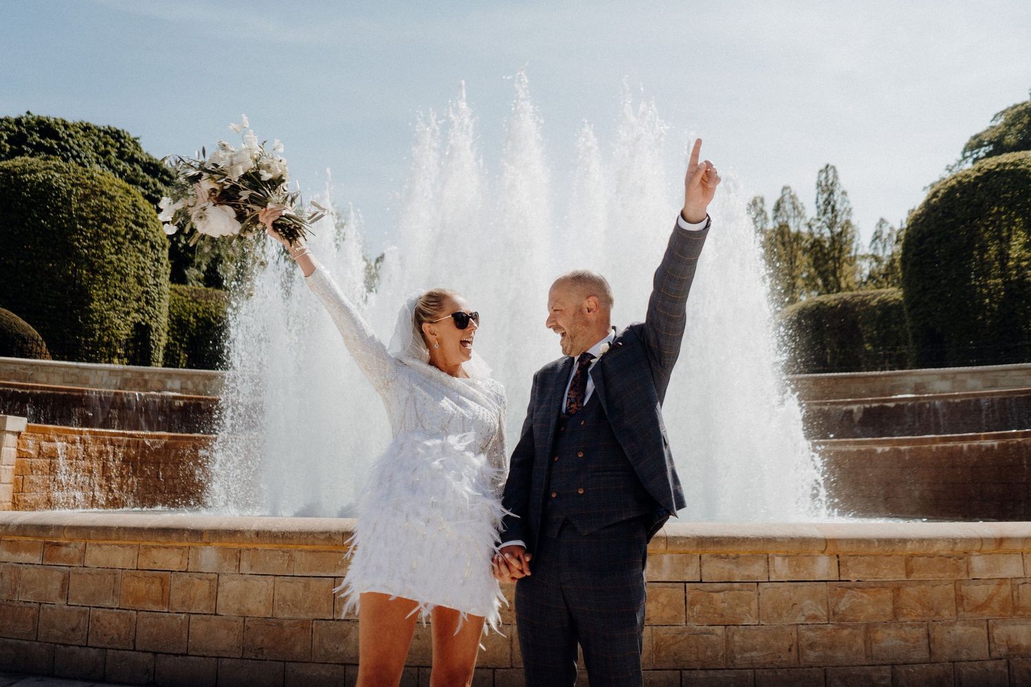 A wedding couple poses by a stone wall with water fountains spraying in the background during their outdoor celebration.