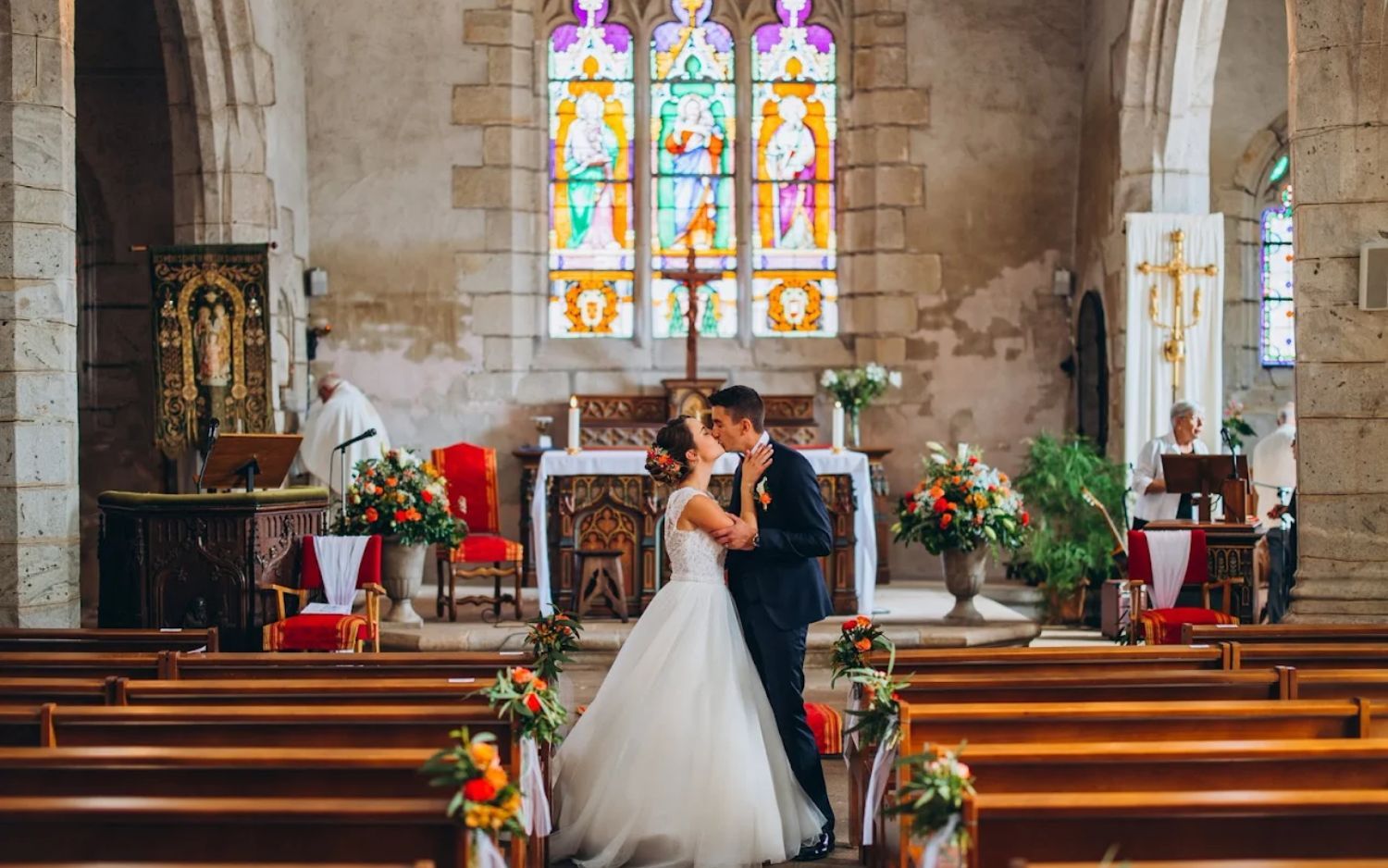 A wedding ceremony inside a historic church with stained glass windows and wooden pews decorated with floral arrangements.