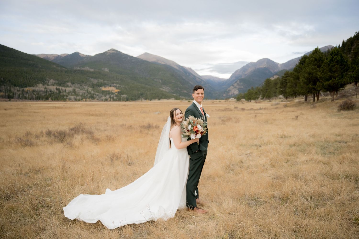 Couple in wedding attire embracing in a scenic mountain meadow with golden grass and mountain peaks in the distance.