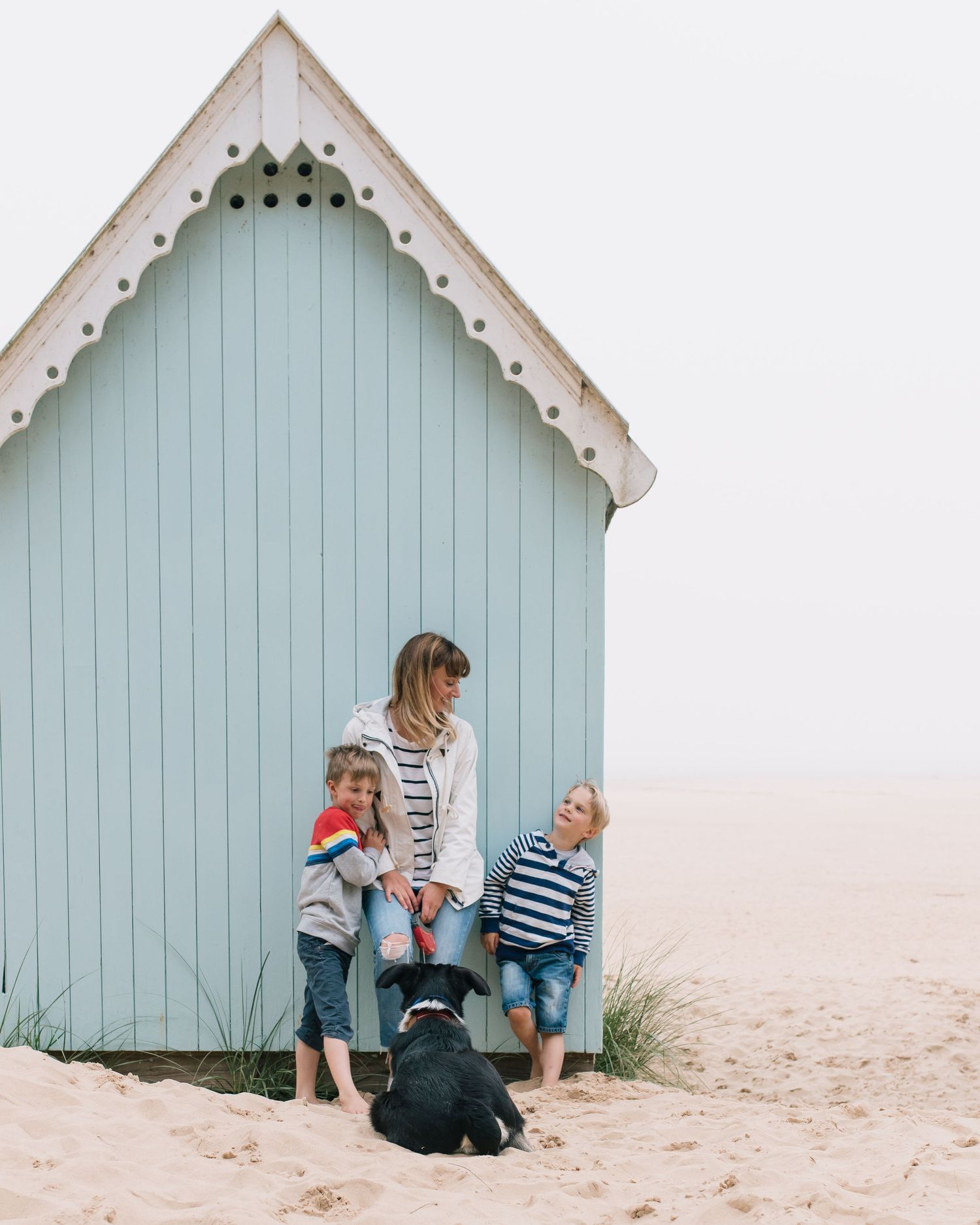 Pale blue beach hut with decorative white trim stands against sandy dunes at the seashore.