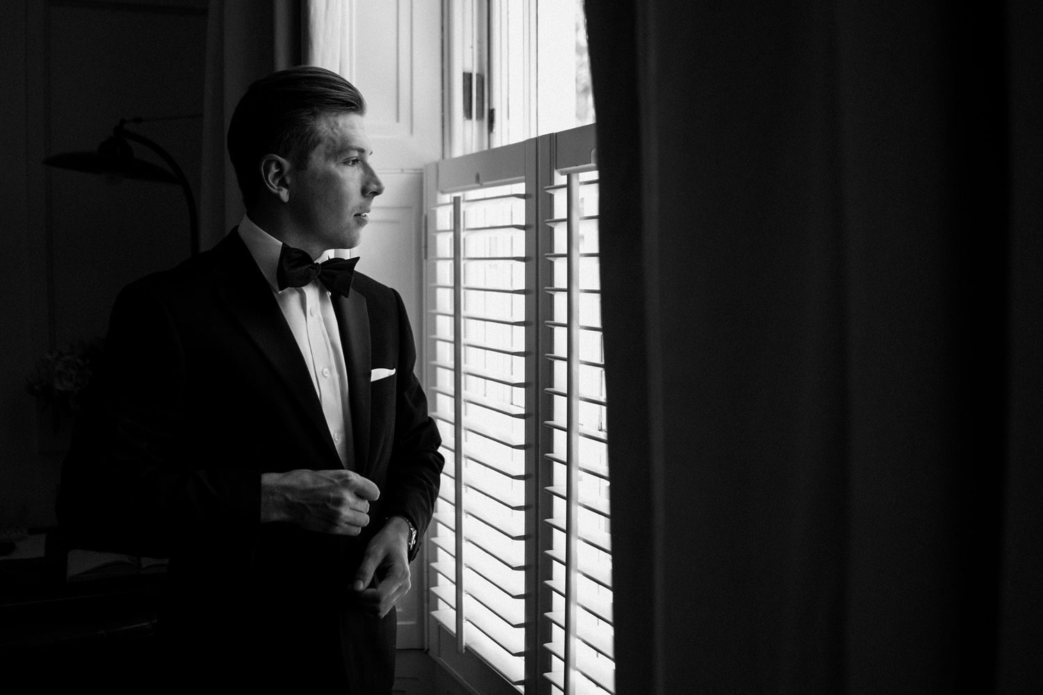 A man in a formal tuxedo and bowtie gazes thoughtfully through window blinds in dramatic black and white lighting in Nova Scotia.