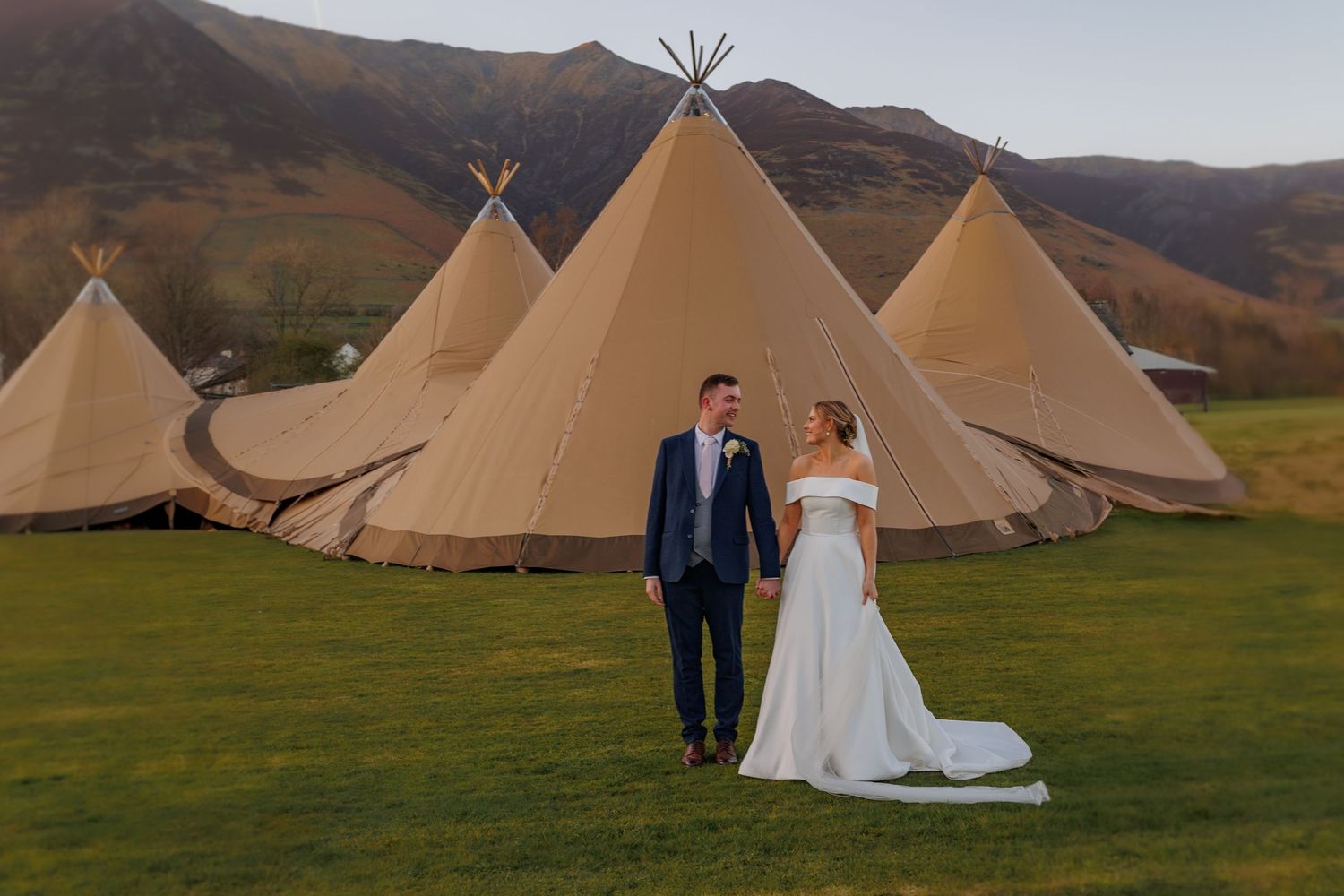 A couple shares an intimate moment in their wedding attire by evergreen trees on a sunny outdoor lawn.