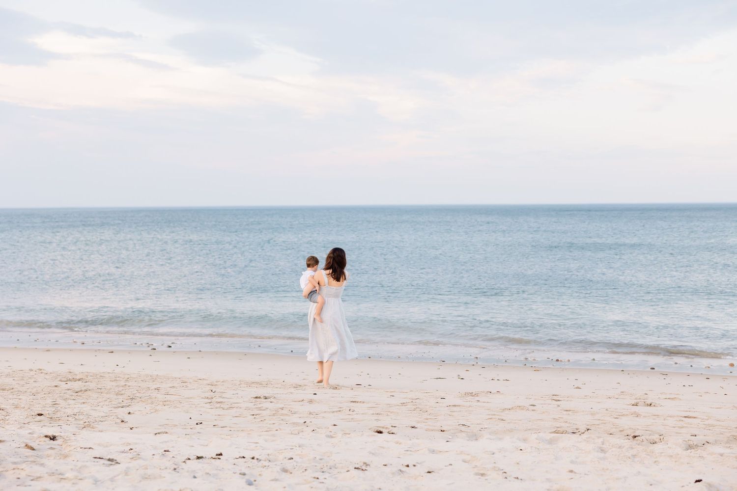 A person in a white dress stands alone on a sandy beach looking out at the calm blue ocean horizon.
