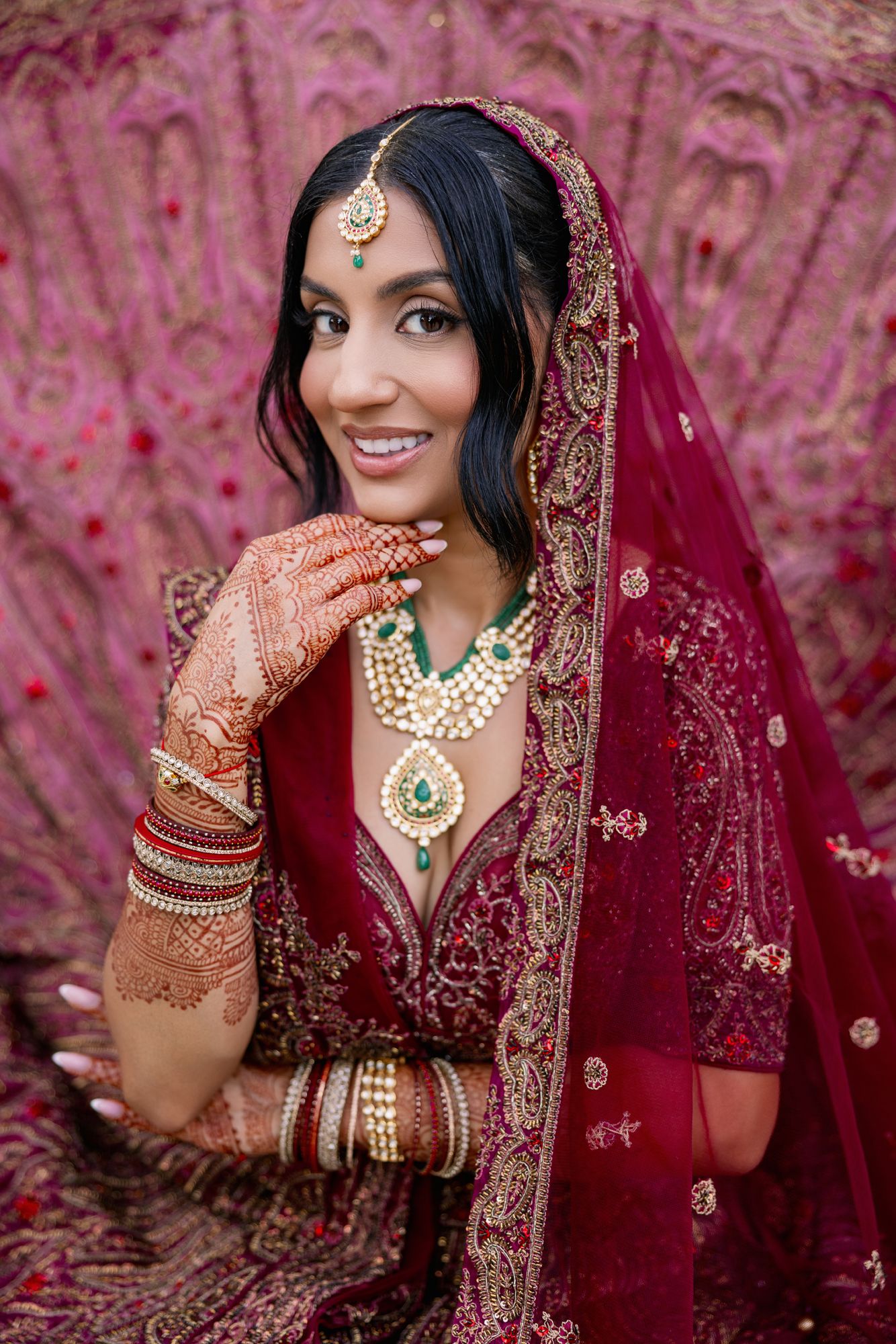 Traditional Indian bride wearing burgundy lehenga with intricate embroidery, henna designs and ornate jewelry against pink backdrop.