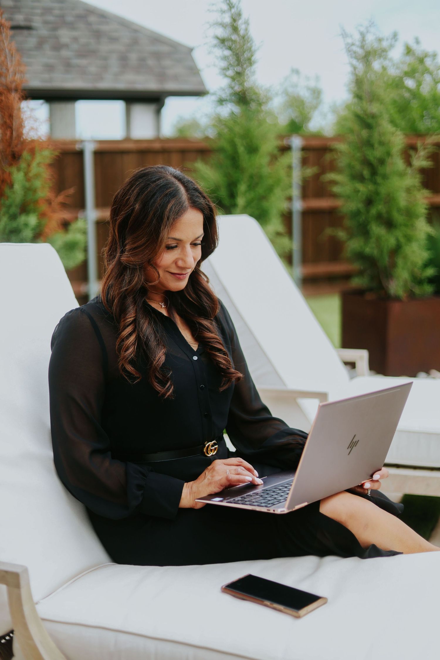 Someone works on a laptop while relaxing on a white outdoor lounge chair with greenery in the background.