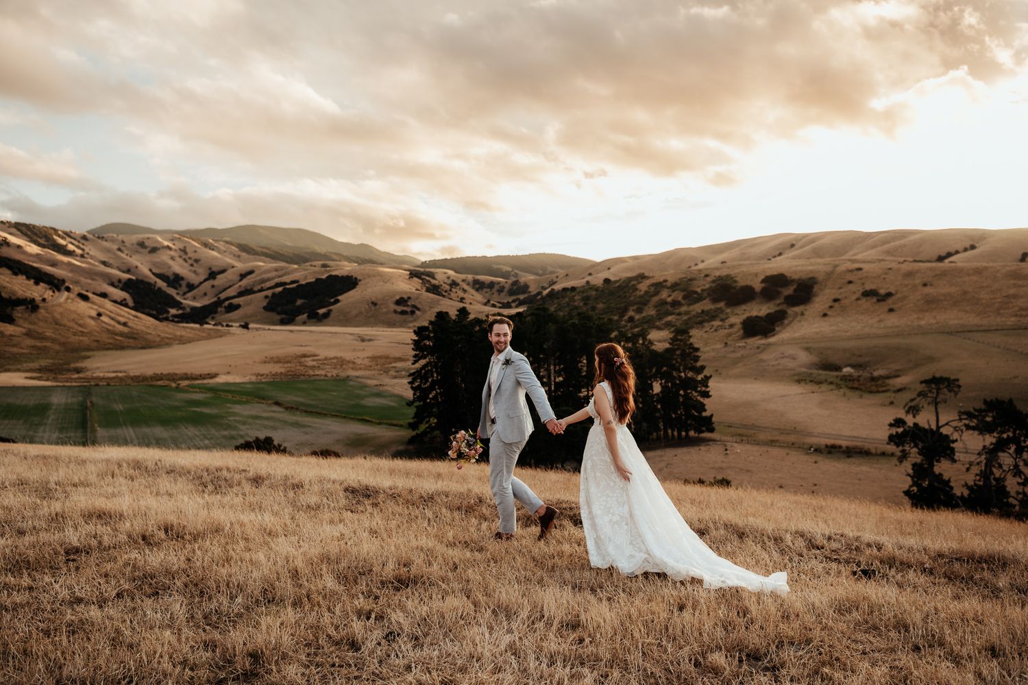 A couple in wedding attire walk hand in hand across a golden hillside at sunset at Tironui Farm Martinborough by August and Vine