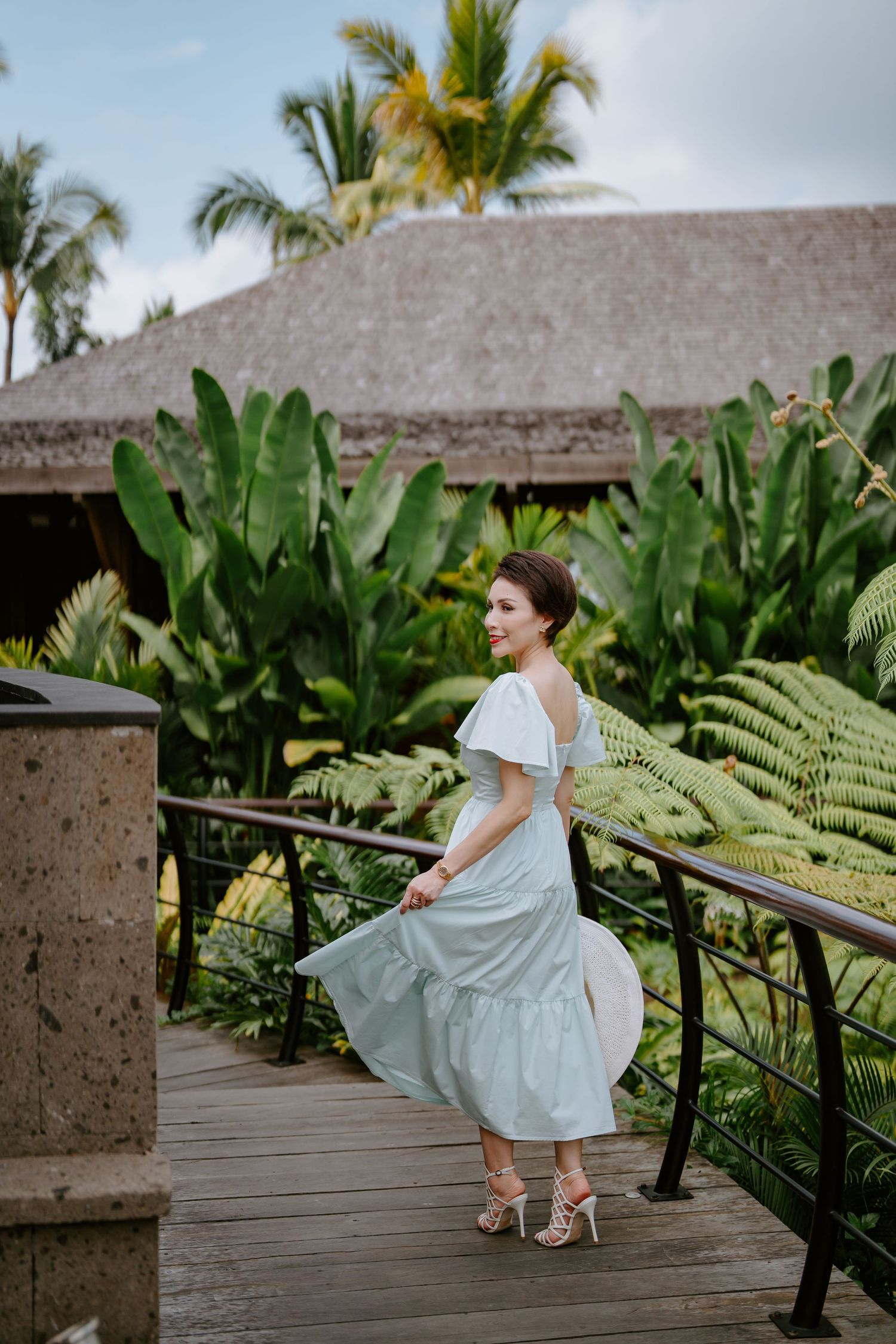 A flowing dress catches movement on a wooden walkway with tropical palms and thatched roof backdrop.