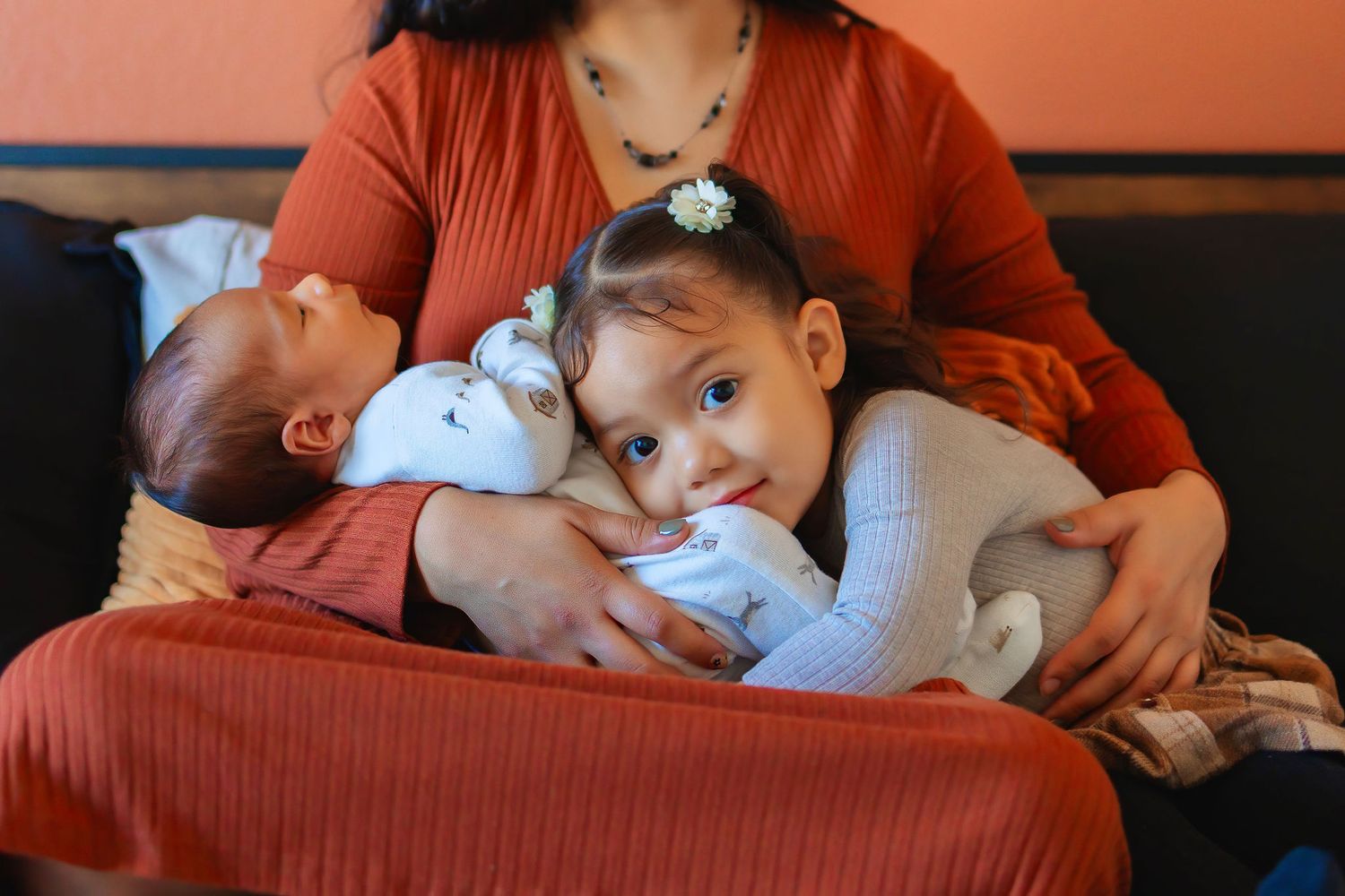 A mother in an orange top holds a toddler girl and newborn baby on her lap, the girl cuddling the infant tenderly.