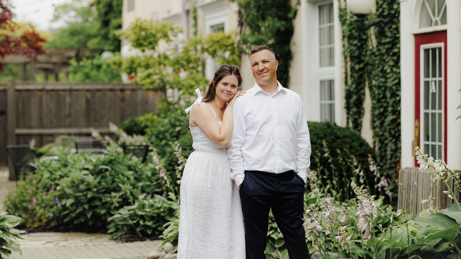 Couple stands together on garden patio surrounded by lush greenery and white flowers next to historic house.