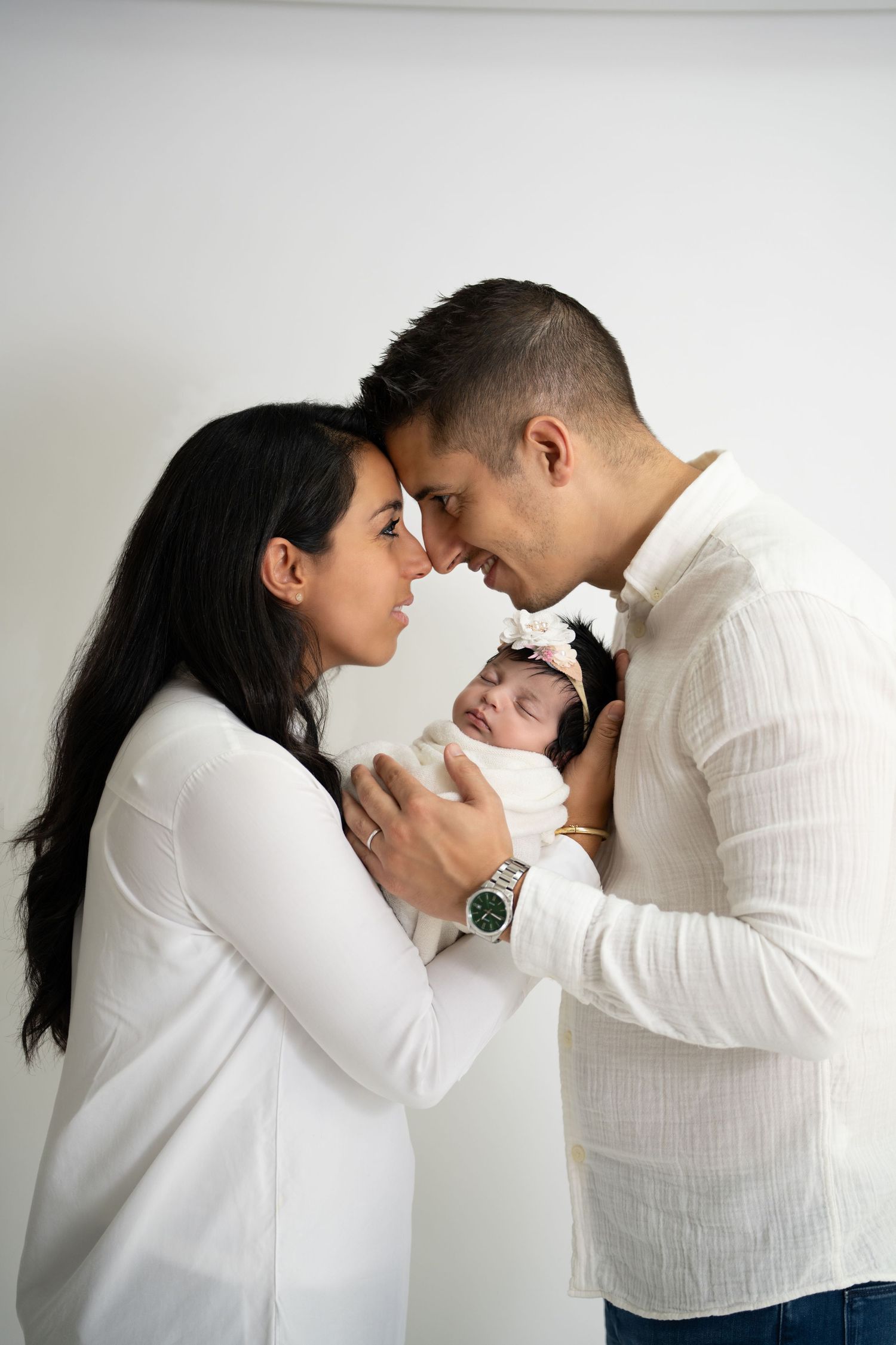 A tender moment as loving parents in white outfits hold their sleeping newborn baby between them against a light background.