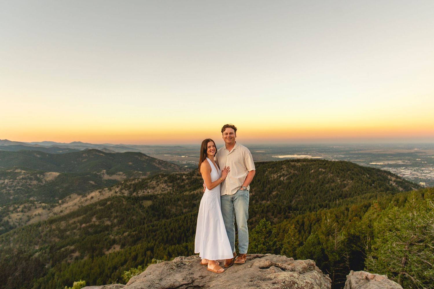 A couple stands on a mountain summit at sunset overlooking a scenic valley and rolling hills in the distance.