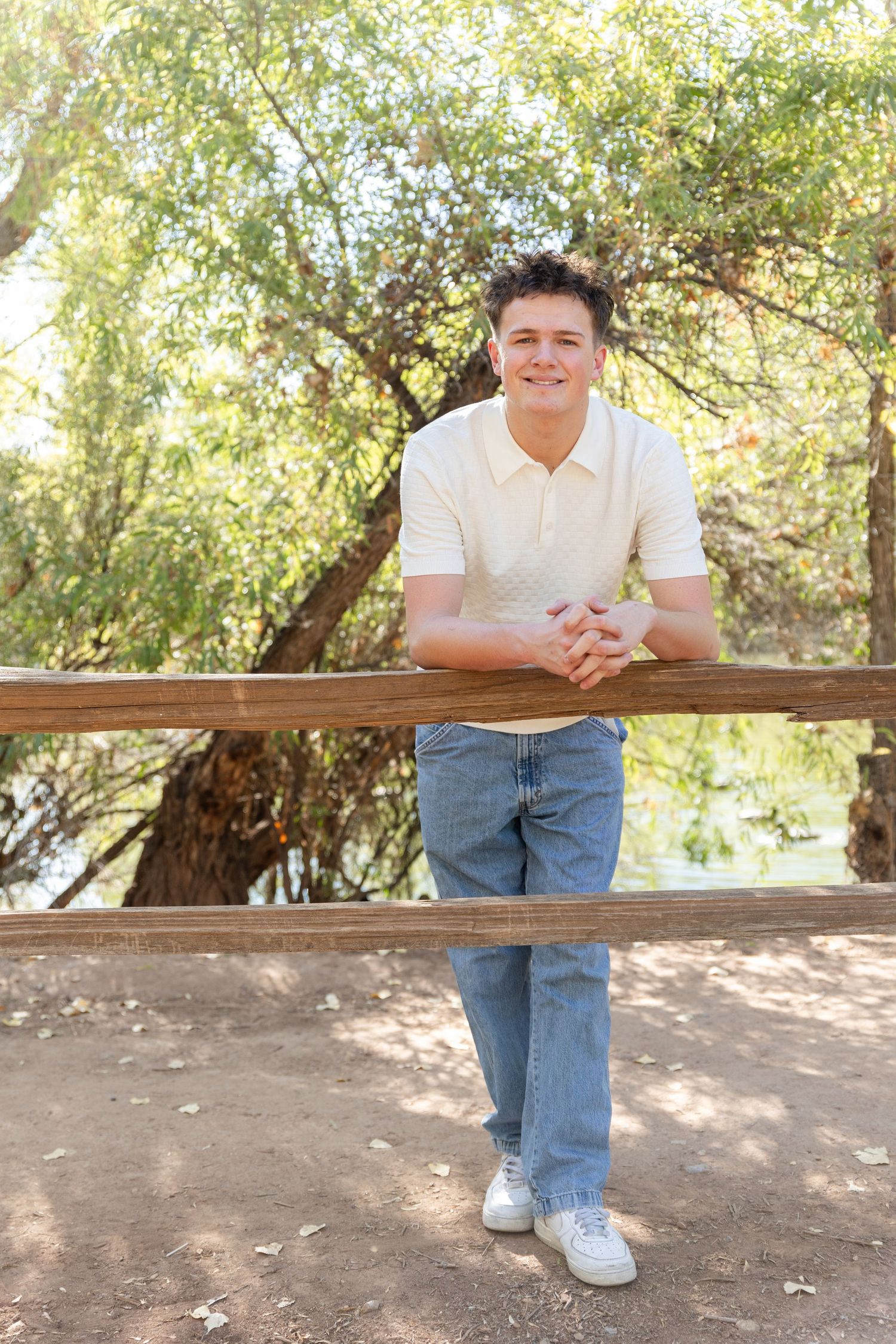 senior boy leaning with forearms against a fence with a lake in and trees in background