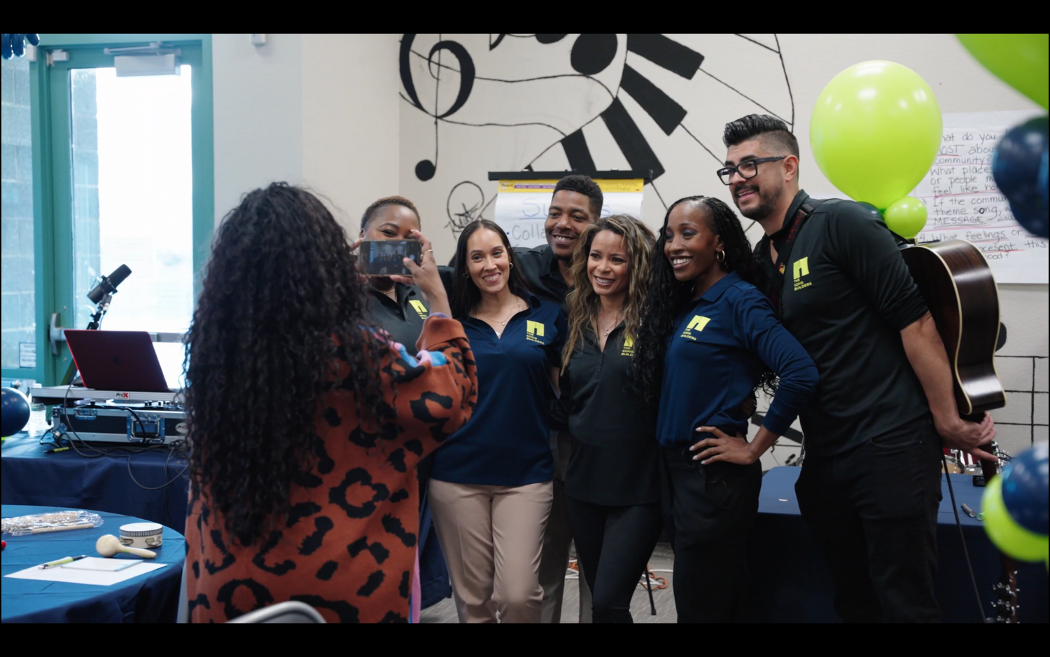Group of people gathered at an indoor event with green balloons visible in the background.