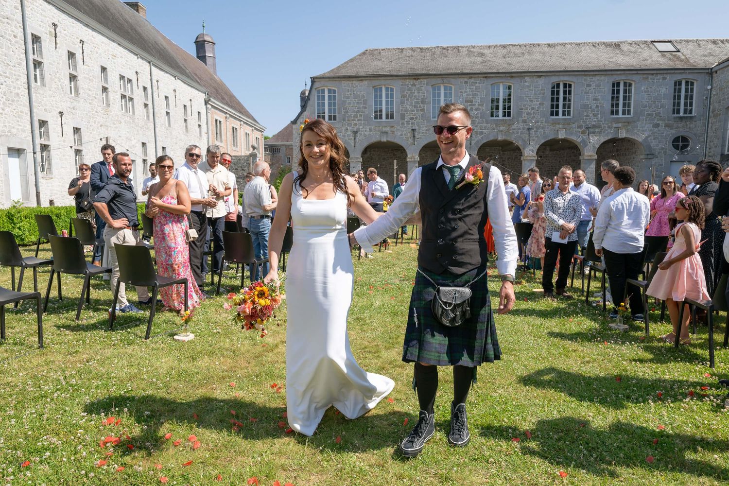 Wedding celebration in front of historic stone building with guest in traditional Scottish kilt attire and formal dress.