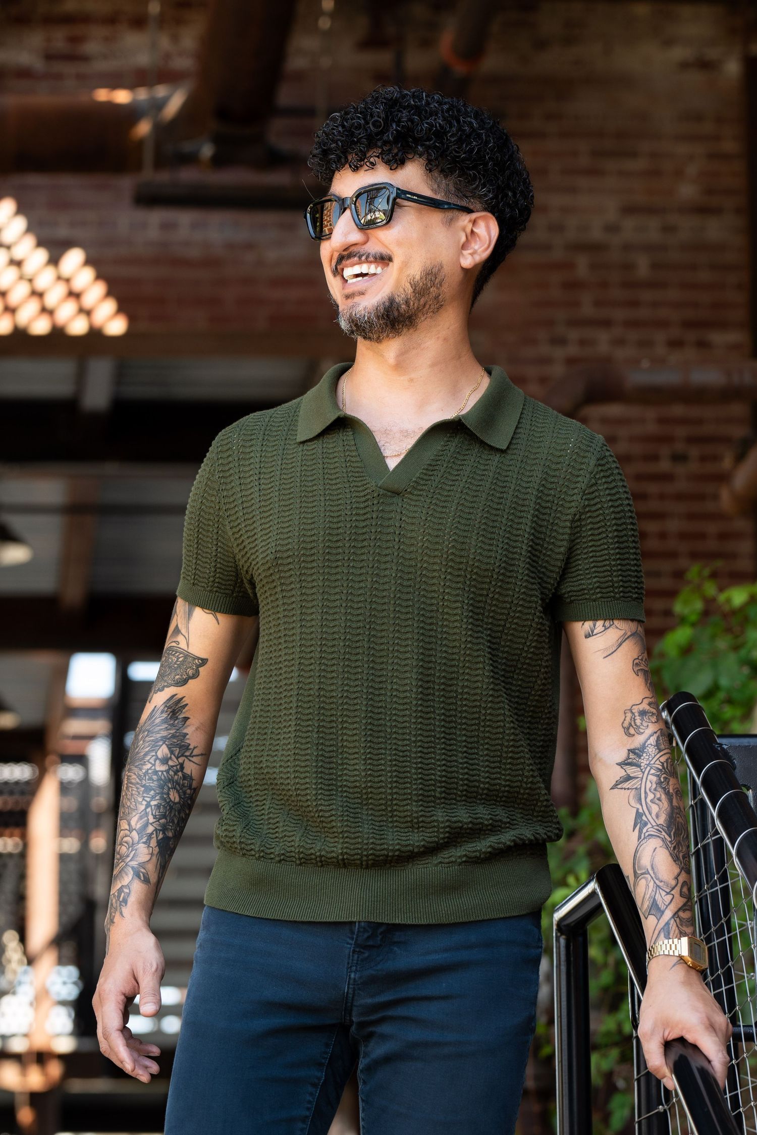 Person wearing olive green knit polo and sunglasses smiles while standing near brick wall with tattoo-covered arms.