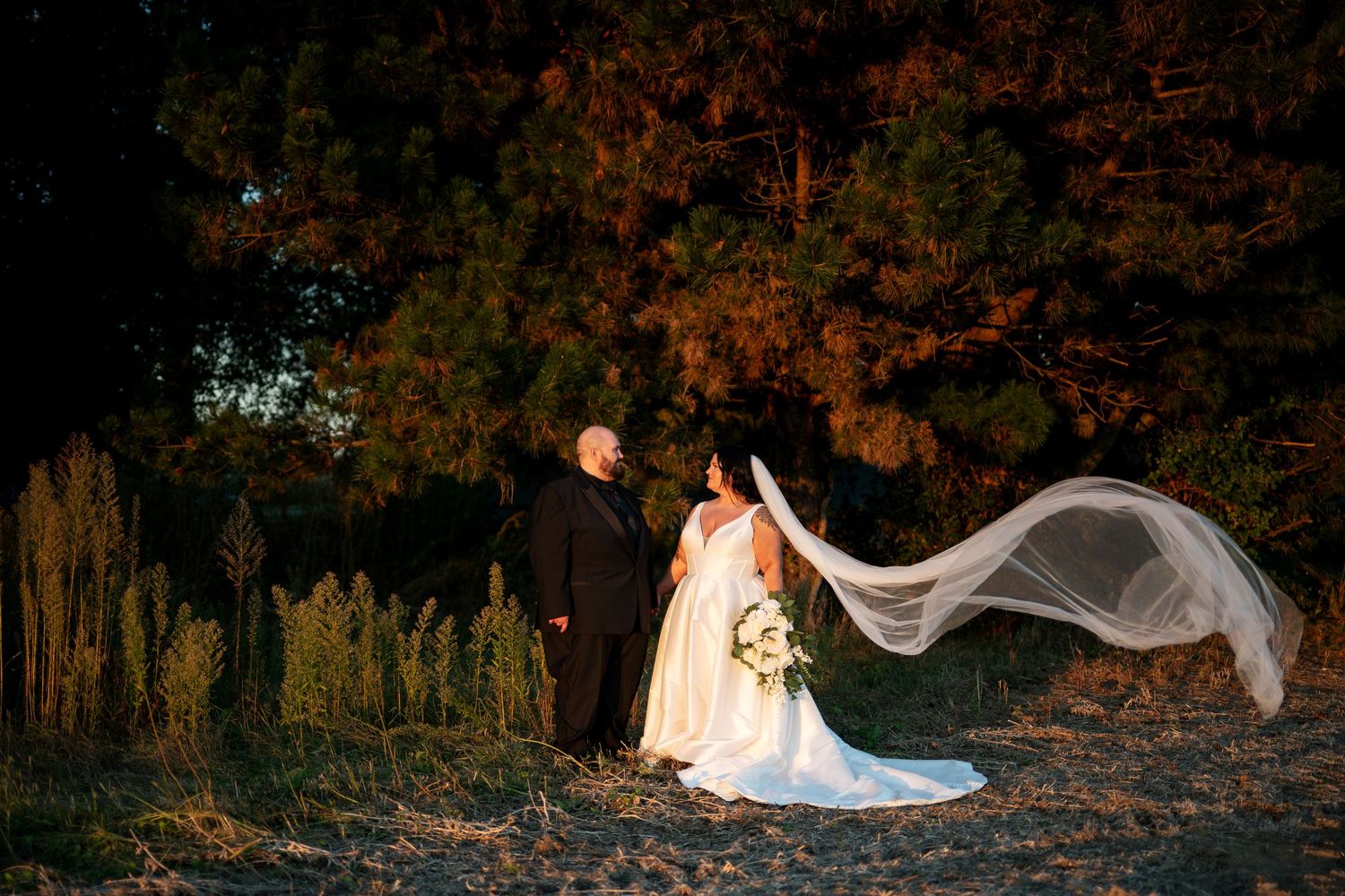 Bride and groom stand together outdoors at golden hour, veil flowing dramatically in the wind.