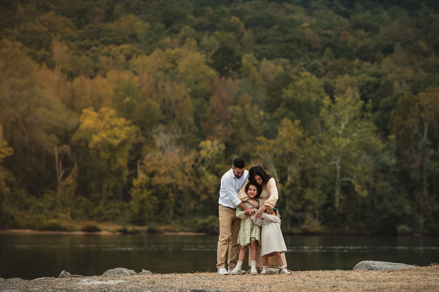 A family stands together on rocky outcrop overlooking a serene lake with autumn forest in background.