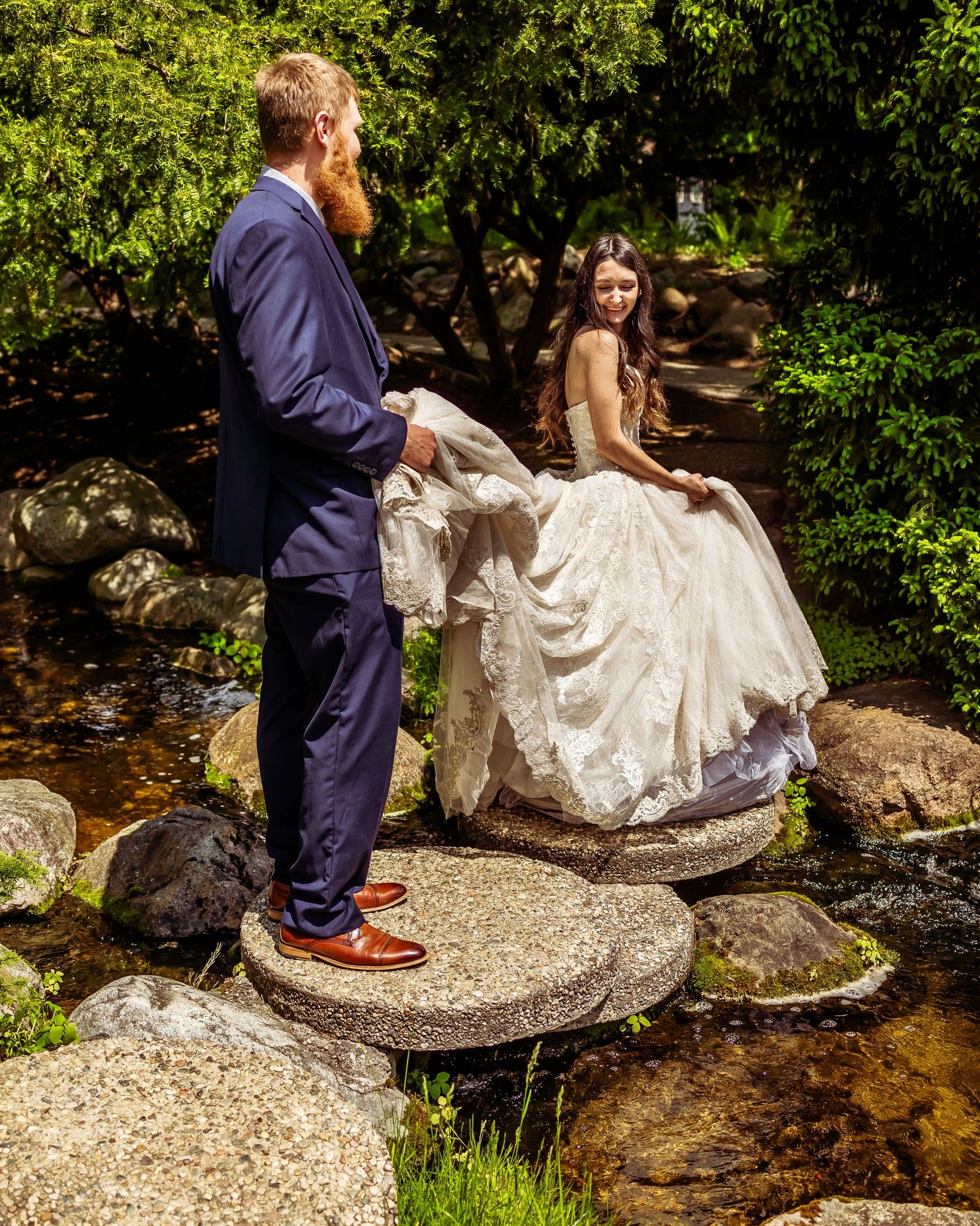 Wedding couple posing on rocks by a stream in a lush garden setting.