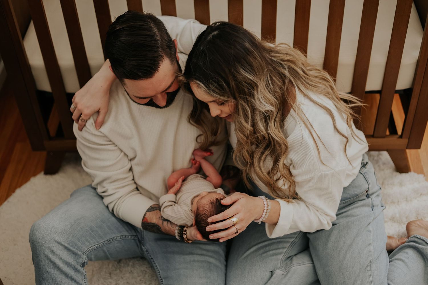 Parents in matching sweaters cuddle their newborn baby on a wooden bench.
