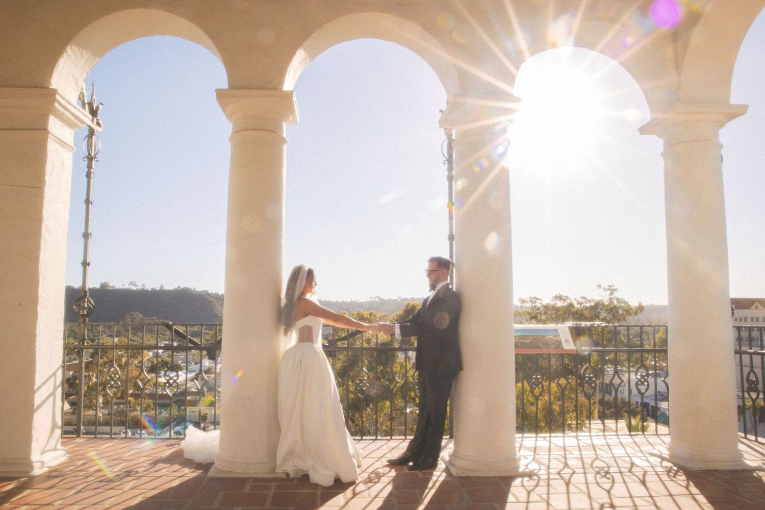 Bride and groom hold hands under sunlit arched columns at an outdoor wedding venue with scenic views.