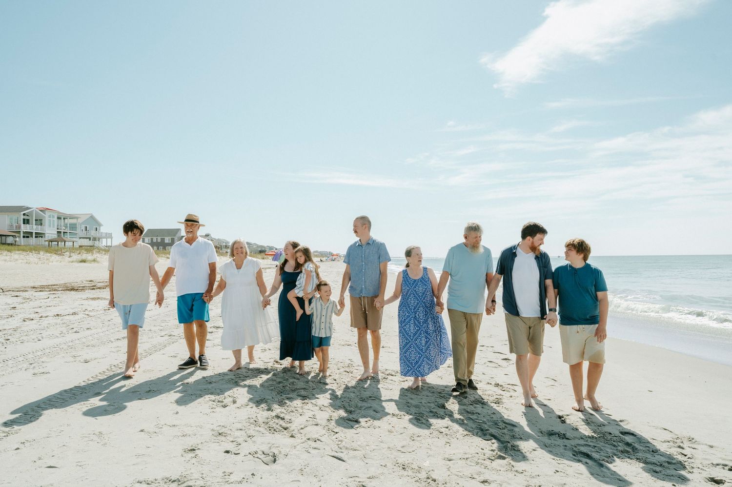 Large family walking hand-in-hand on a sunny beach, dressed in coordinated outfits, with ocean waves and coastal homes in the background.