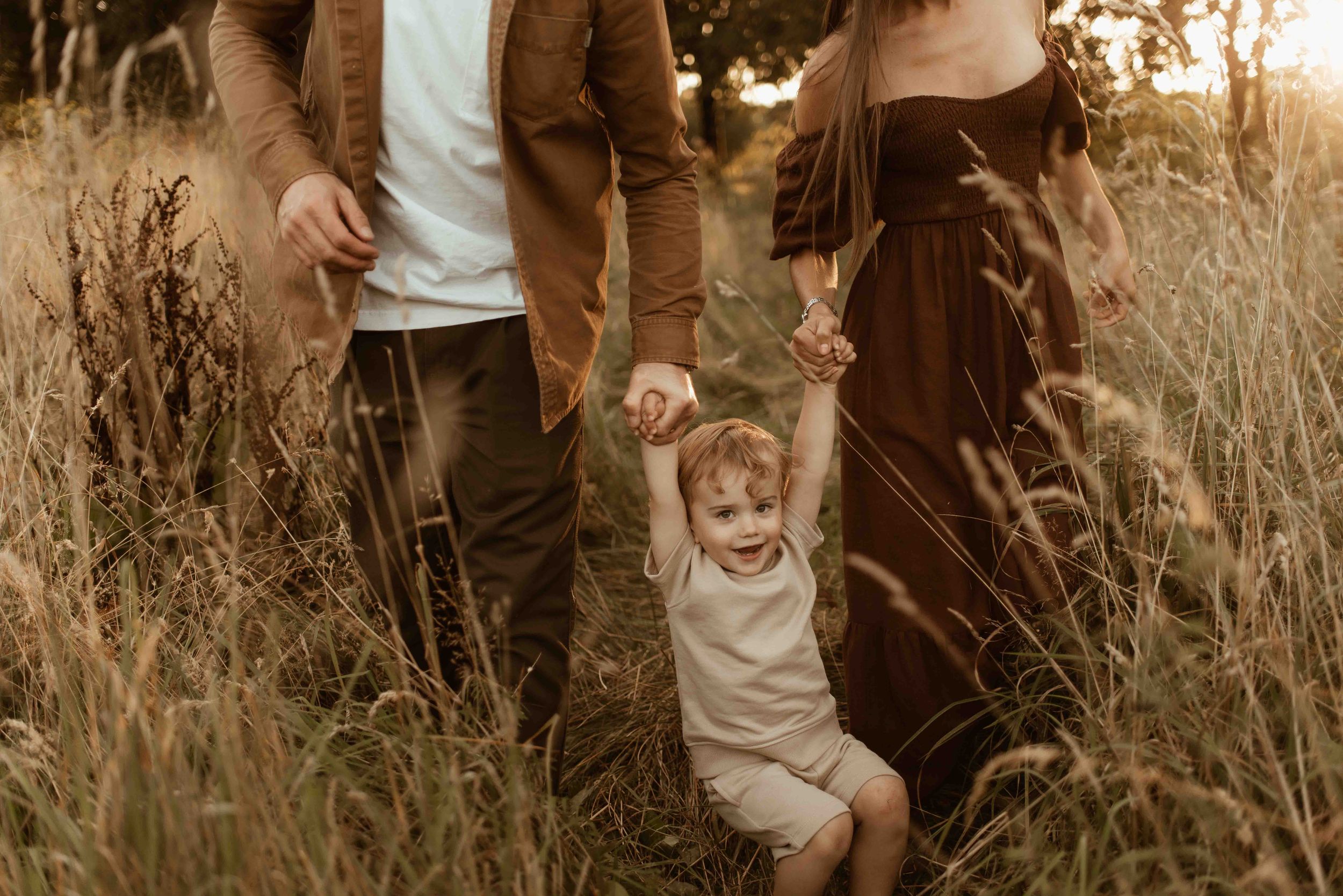 Family walking through golden field at sunset, parents holding toddler's hands as child laughs joyfully.
