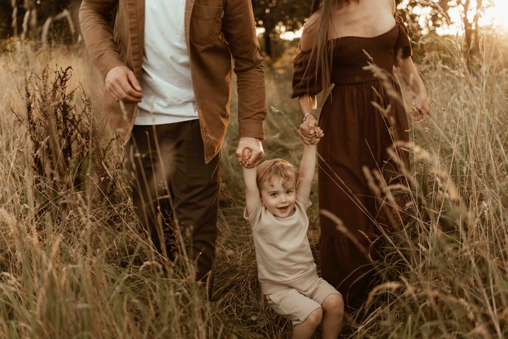 Family walking through golden field at sunset, parents holding toddler's hands as child laughs joyfully.