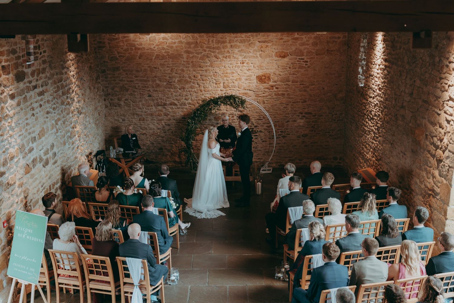 Elegant wedding ceremony in a rustic brick venue with guests seated as couple exchanges vows under a floral arch.