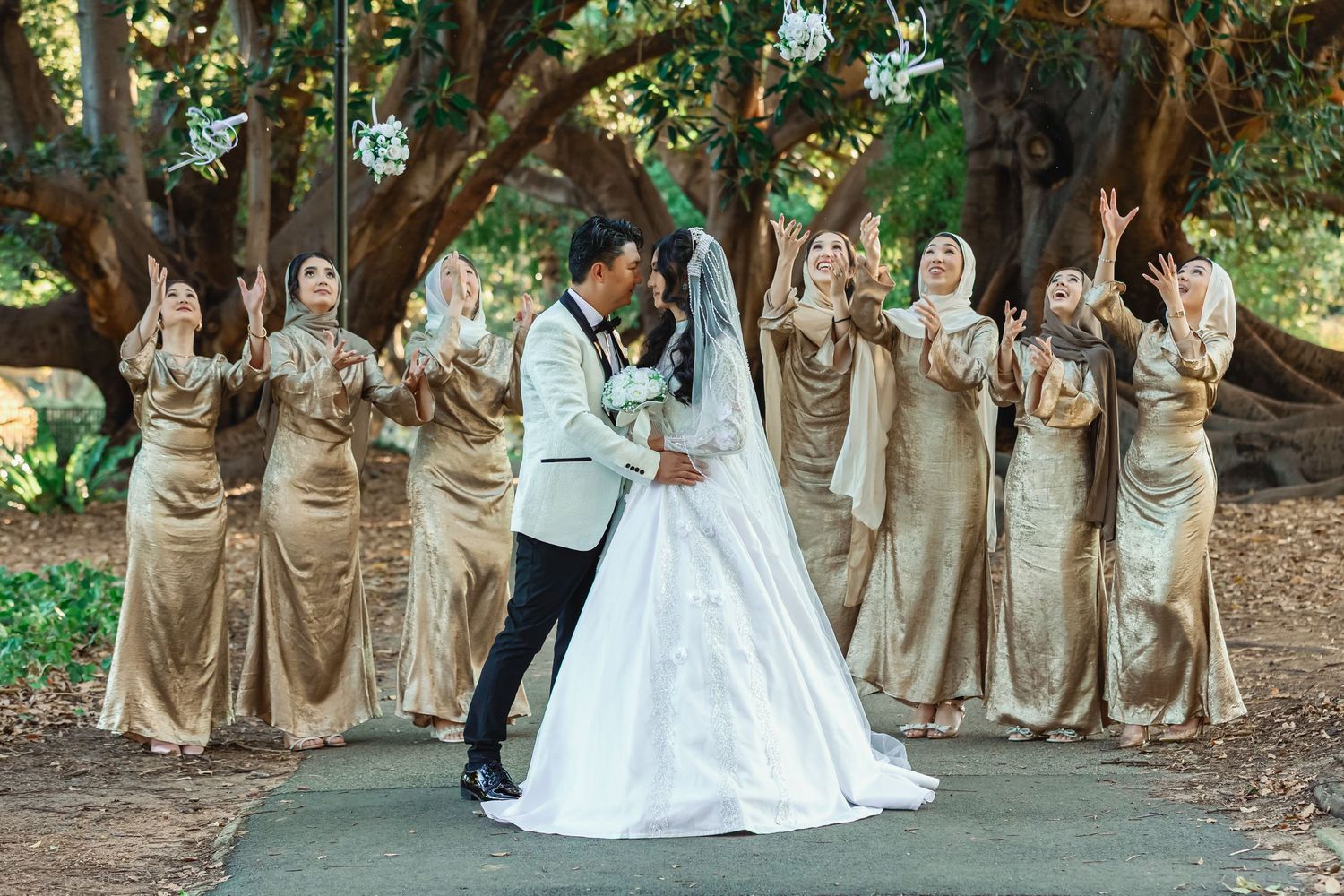 Bride and groom share a romantic moment as bridesmaids in gold gowns raise bouquets beneath a grand tree.