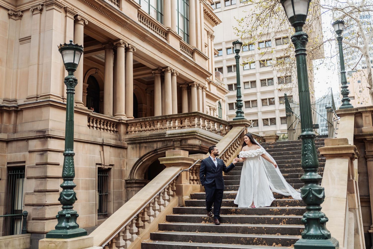 A couple in wedding attire poses on elegant stone steps of a historic building with ornate architecture and lamp posts.