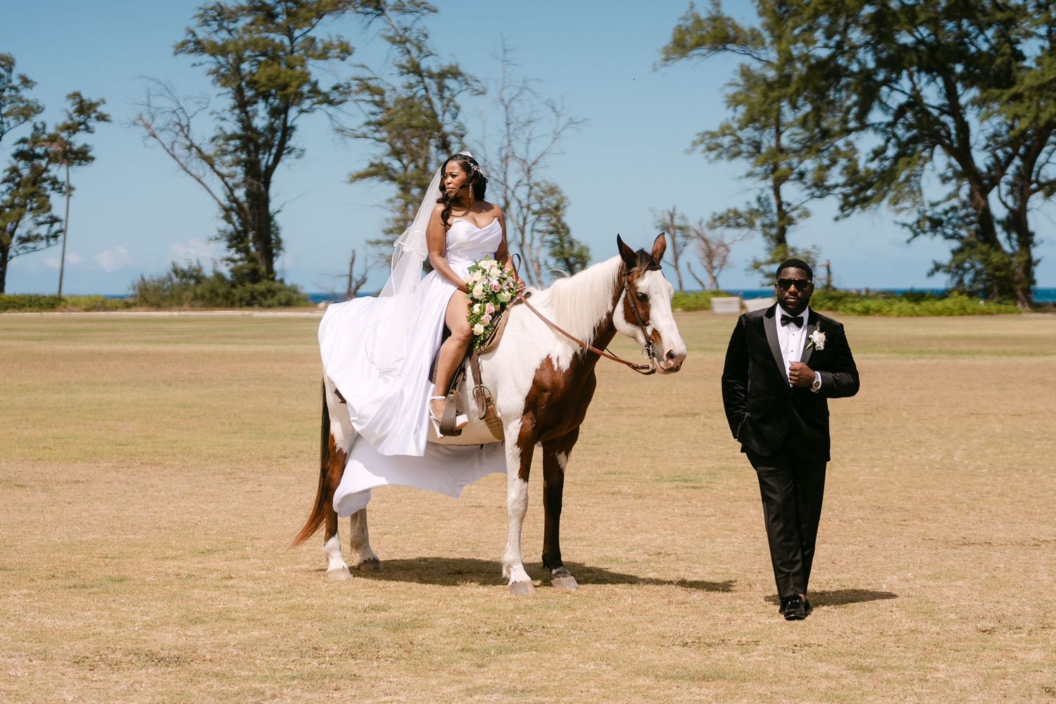 A bride in a white dress sits sidesaddle on a paint horse while someone in a black suit walks beside them in a field.