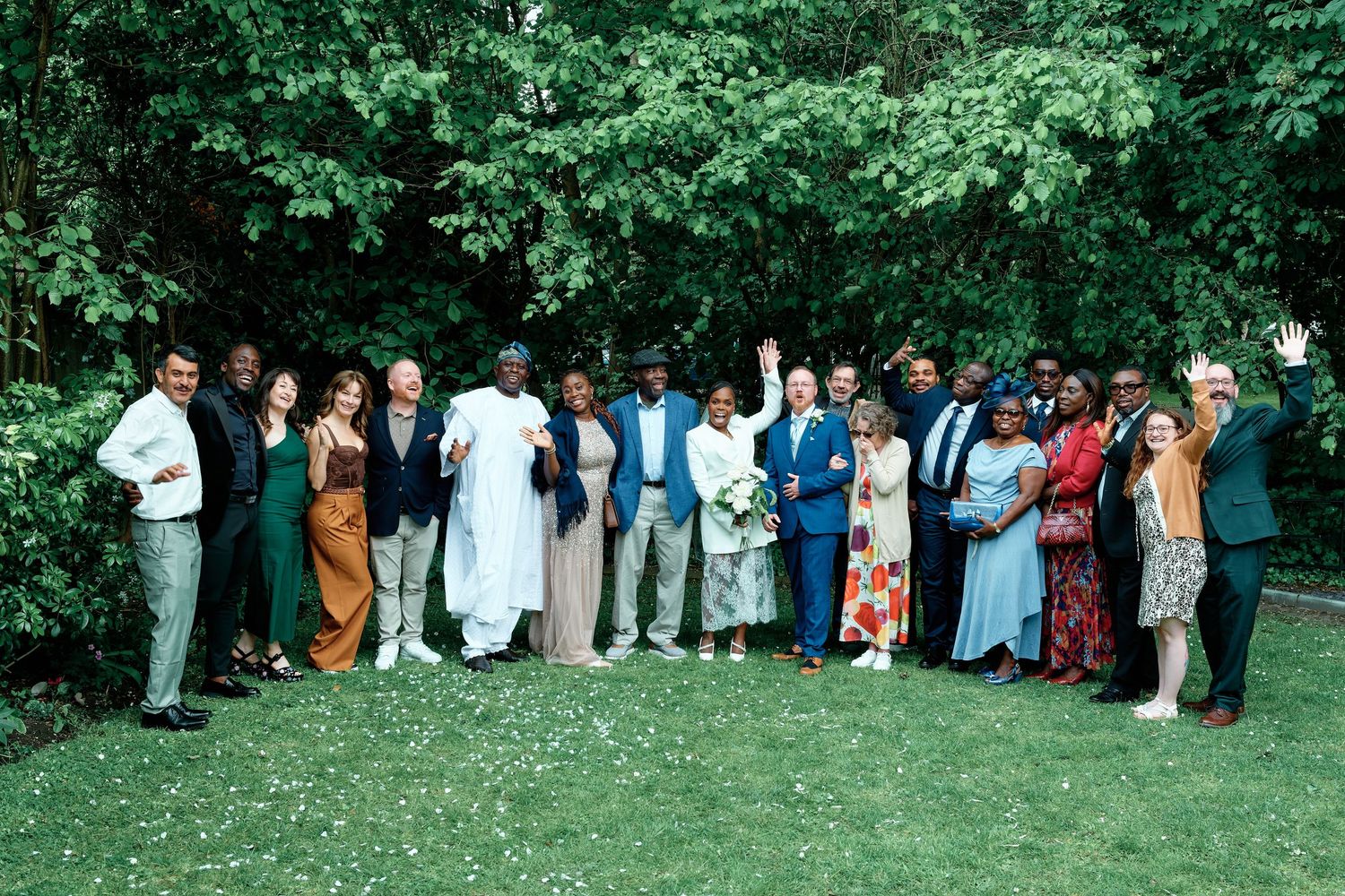 Large diverse group of people posing together outdoors in a lush green garden, waving and smiling at a wedding celebration.