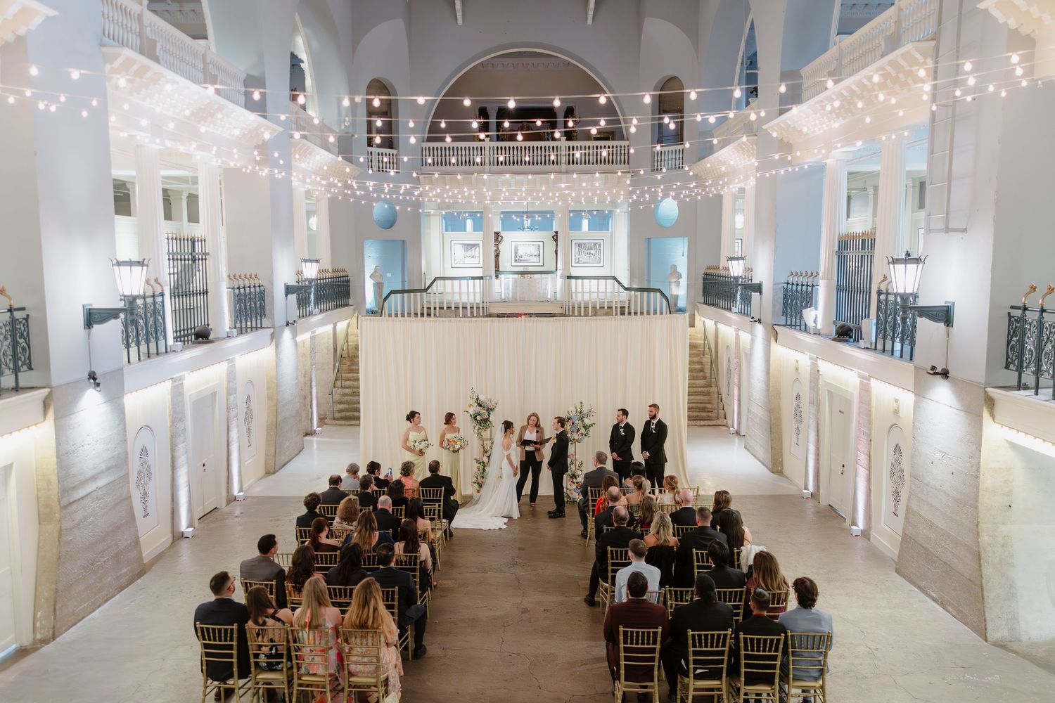 Elegant indoor wedding ceremony in a grand ballroom with string lights, white draping, and guests seated in gold chairs.