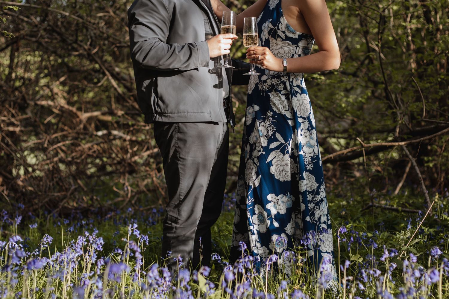 Couple in formal attire toasting with champagne glasses among bluebells in a woodland setting.