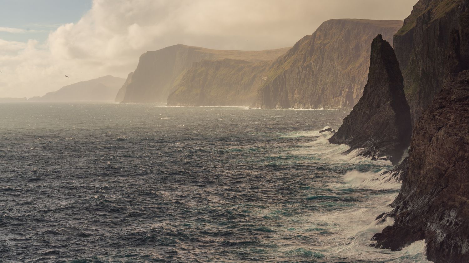 Dramatic ocean cliffs rise from crashing waves under golden stormy light along a rugged coastline.