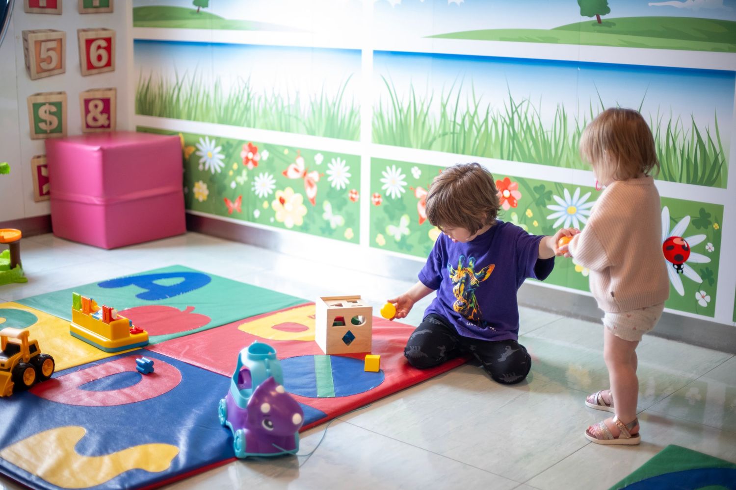 Two toddlers playing with colorful building blocks on an alphabet mat in a bright, cheerful children's playroom.