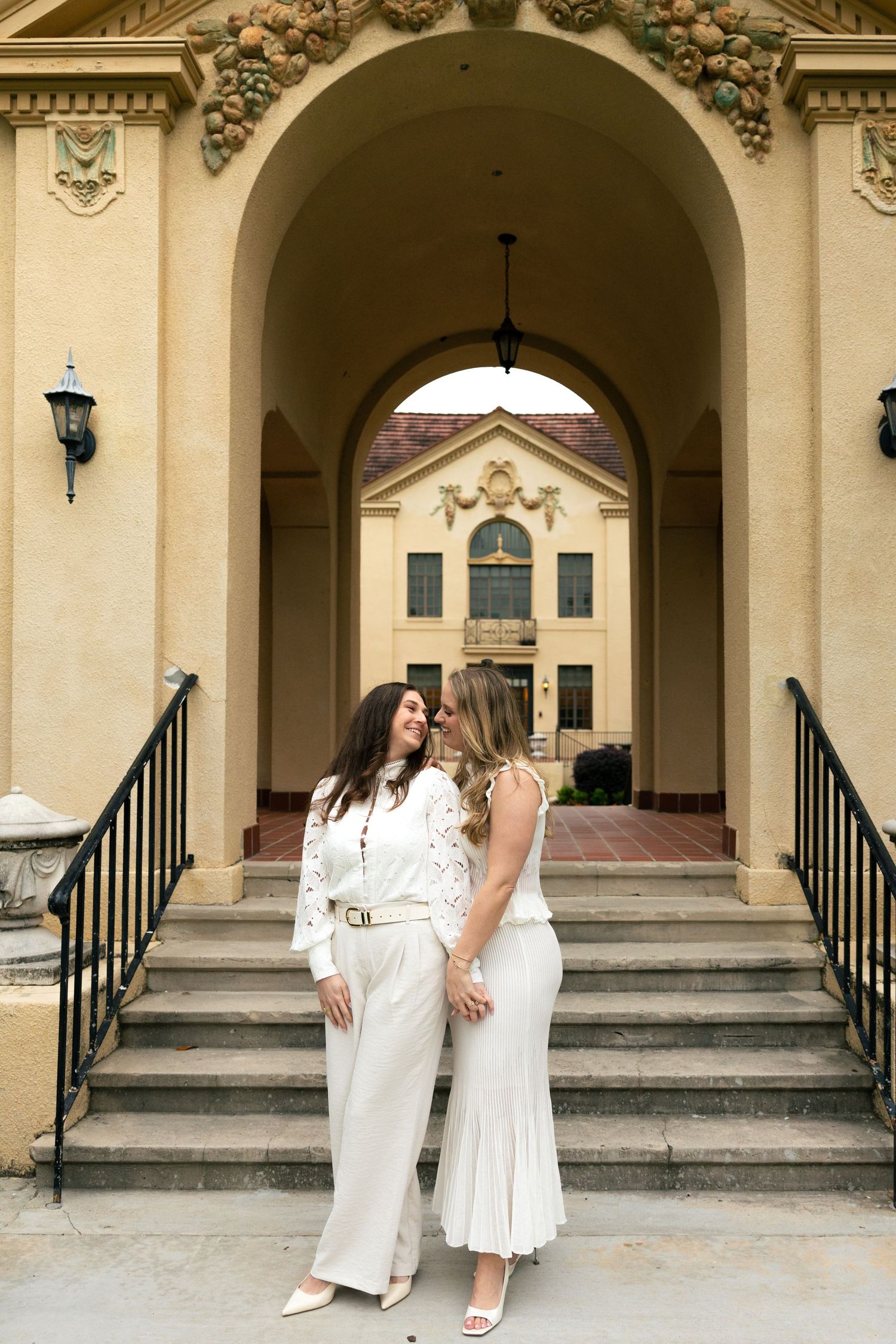 Two women in white outfits share a romantic moment on steps of an elegant building with arched entrance.