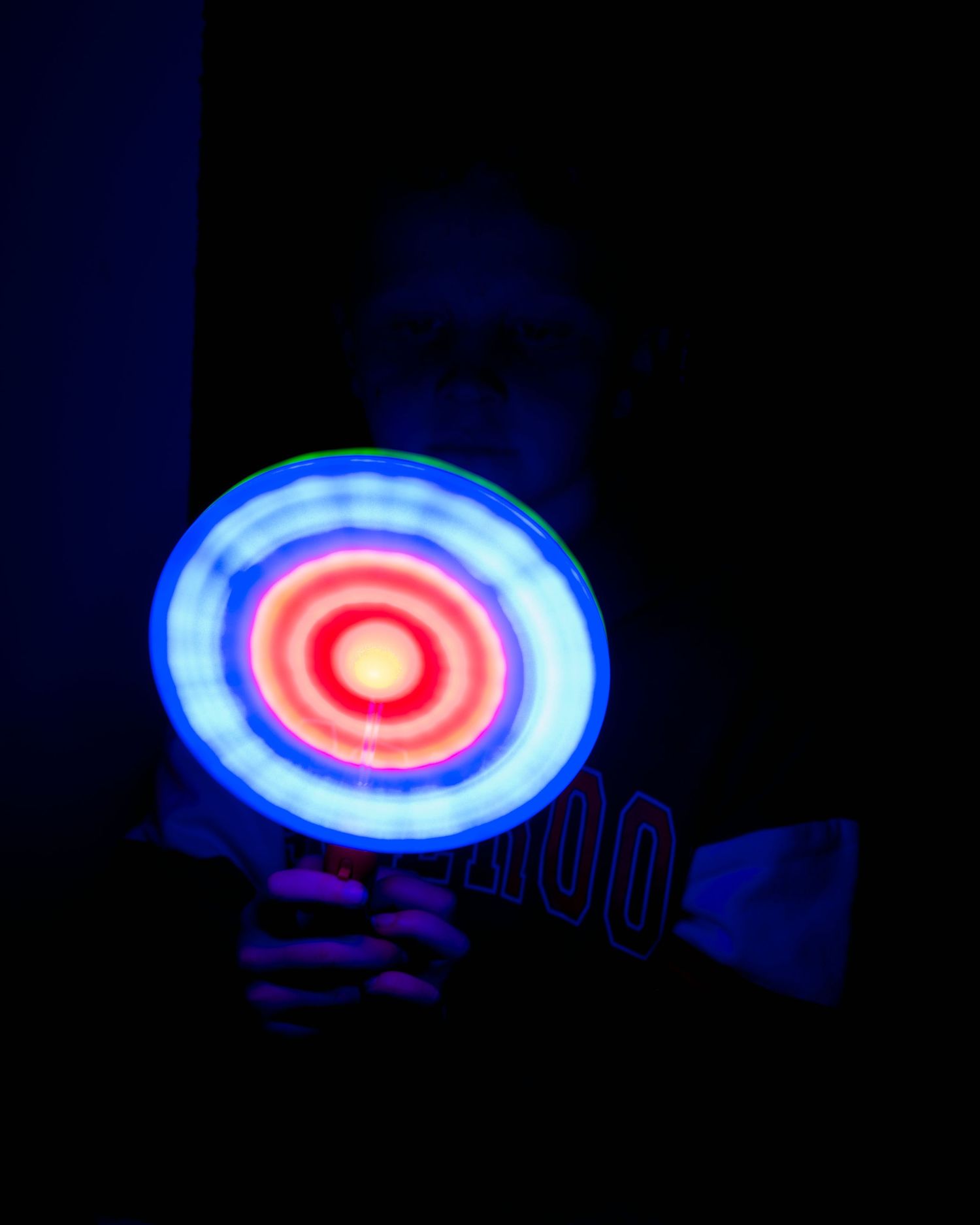 Person holding a glowing neon target disc with concentric red and blue circles in a dark room.
